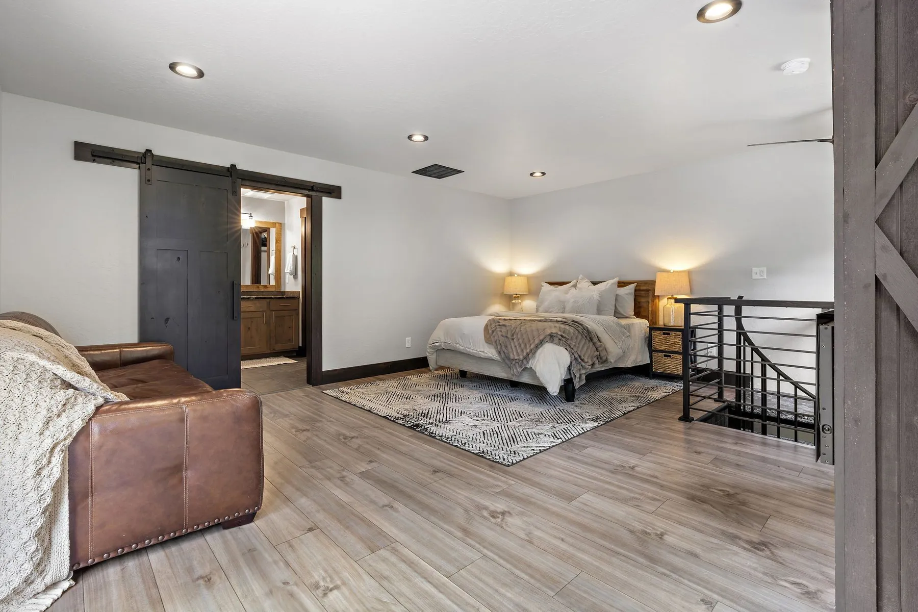 Upstairs primary bedroom with a dark sliding barn door, a black stair railing and light plank flooring.