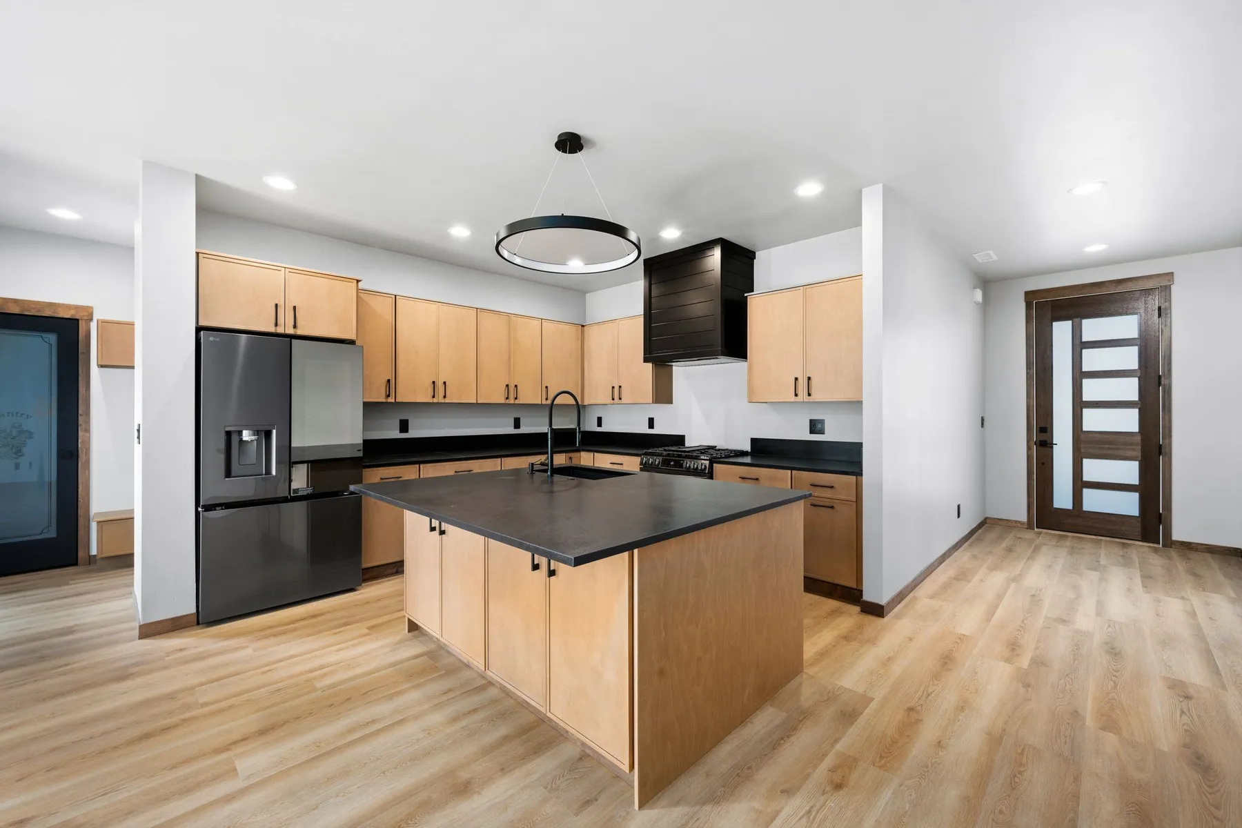 Kitchen with natural maple cabinets, black stone countertops, a black range hood and a ring pendant over the island.