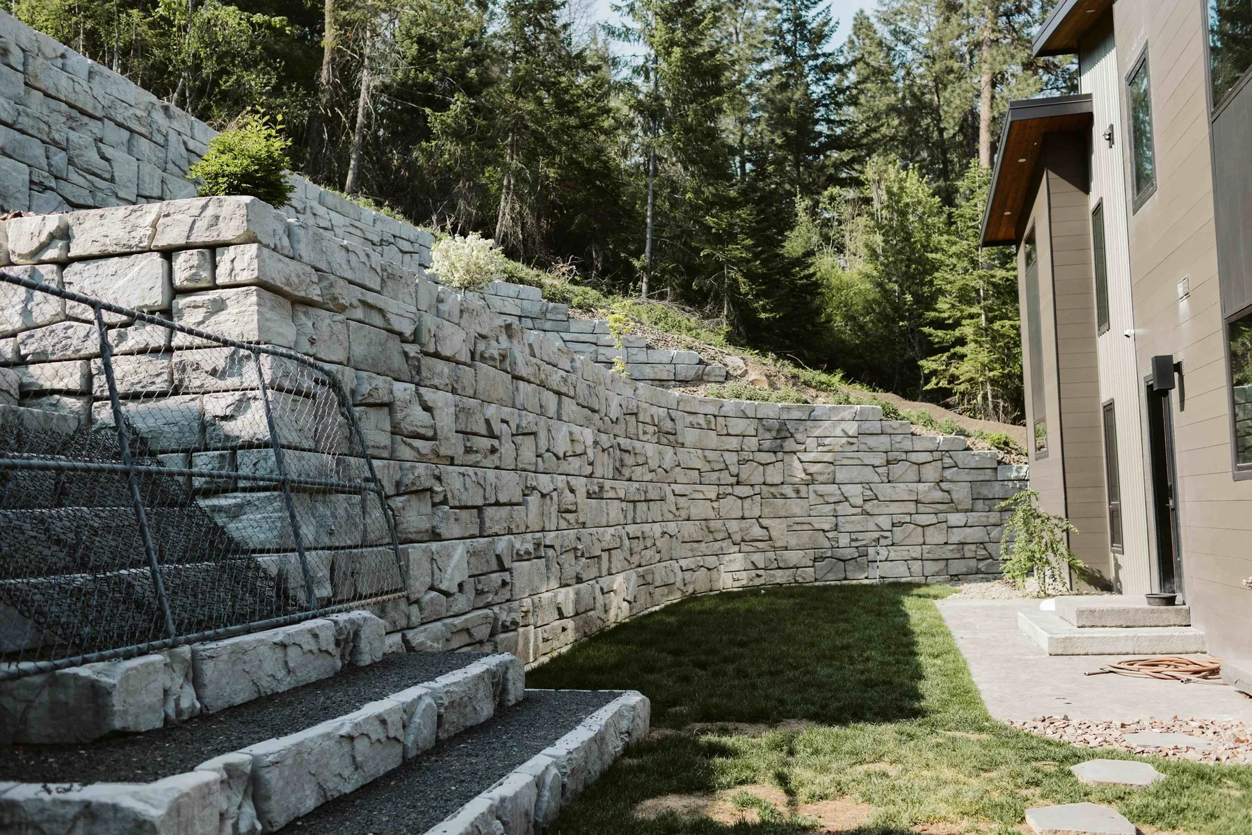 Block retaining wall with built-in stone steps and a railing climbing the slope beside the home.
