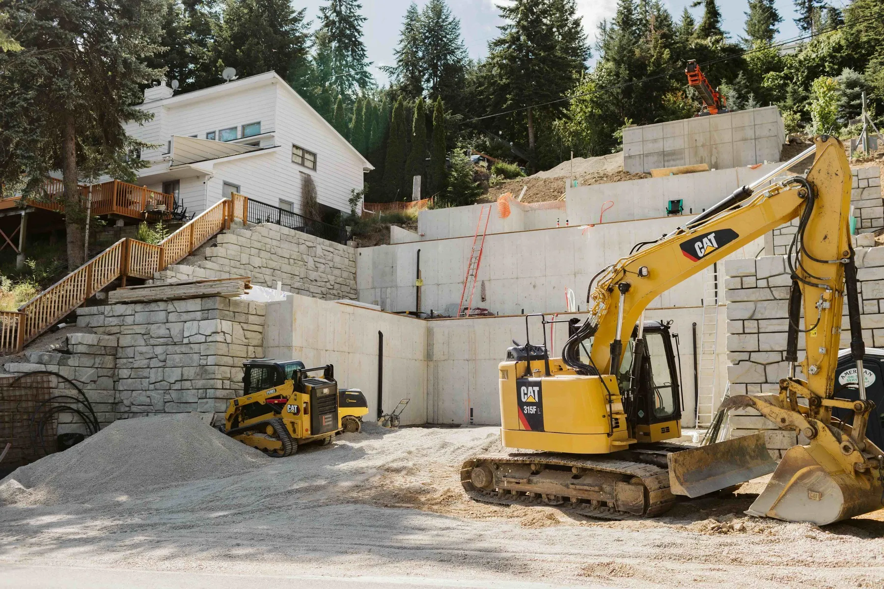 CAT excavator and skid steer working below tiered concrete retaining walls cut into a steep hillside.