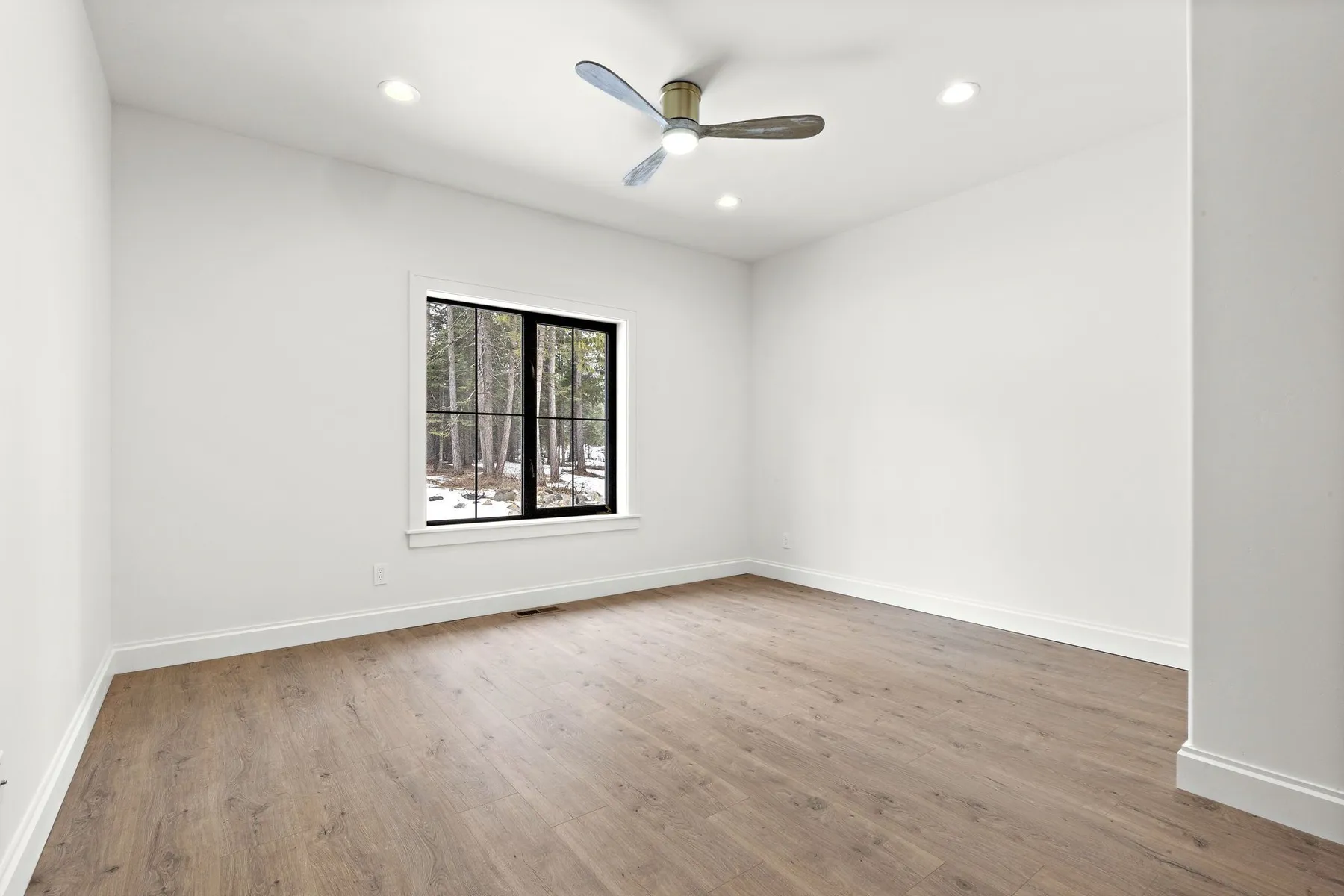 Empty secondary bedroom with light plank flooring, a black-framed window to the trees and a ceiling fan.
