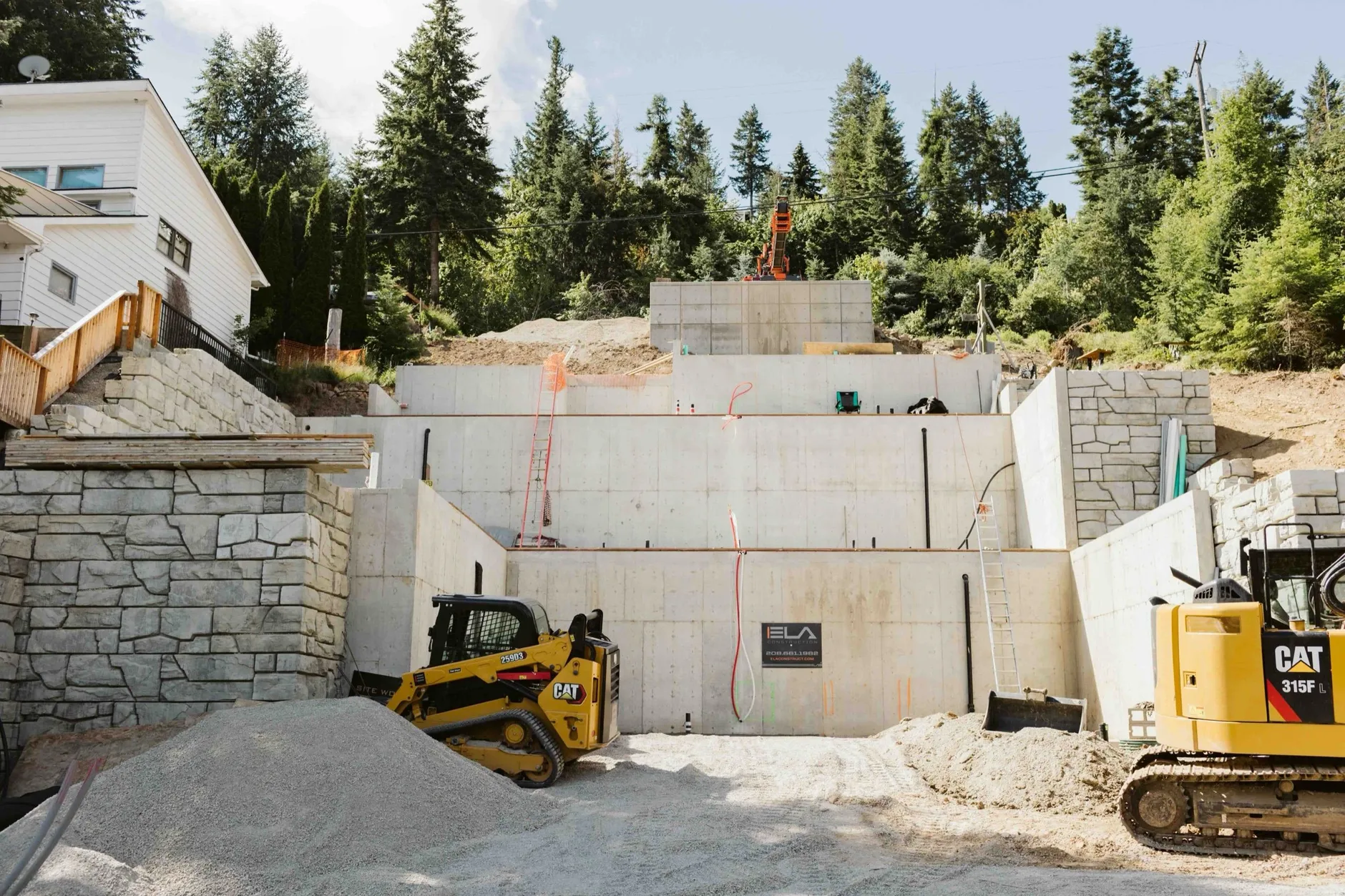 Terraced concrete foundation walls cut into a steep wooded hillside, with a CAT compact track loader and a CAT 315 excavator on the gravel pad below.
