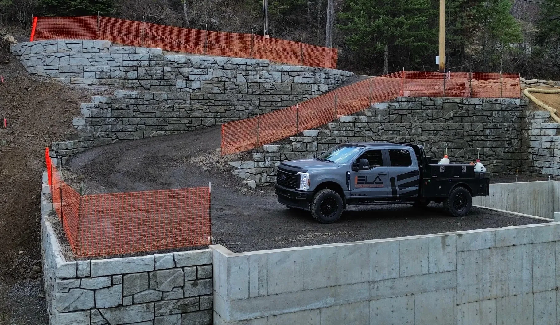 Gray pickup truck with the Ela Construction logo parked on a gravel area near stone retaining walls with orange safety fencing.