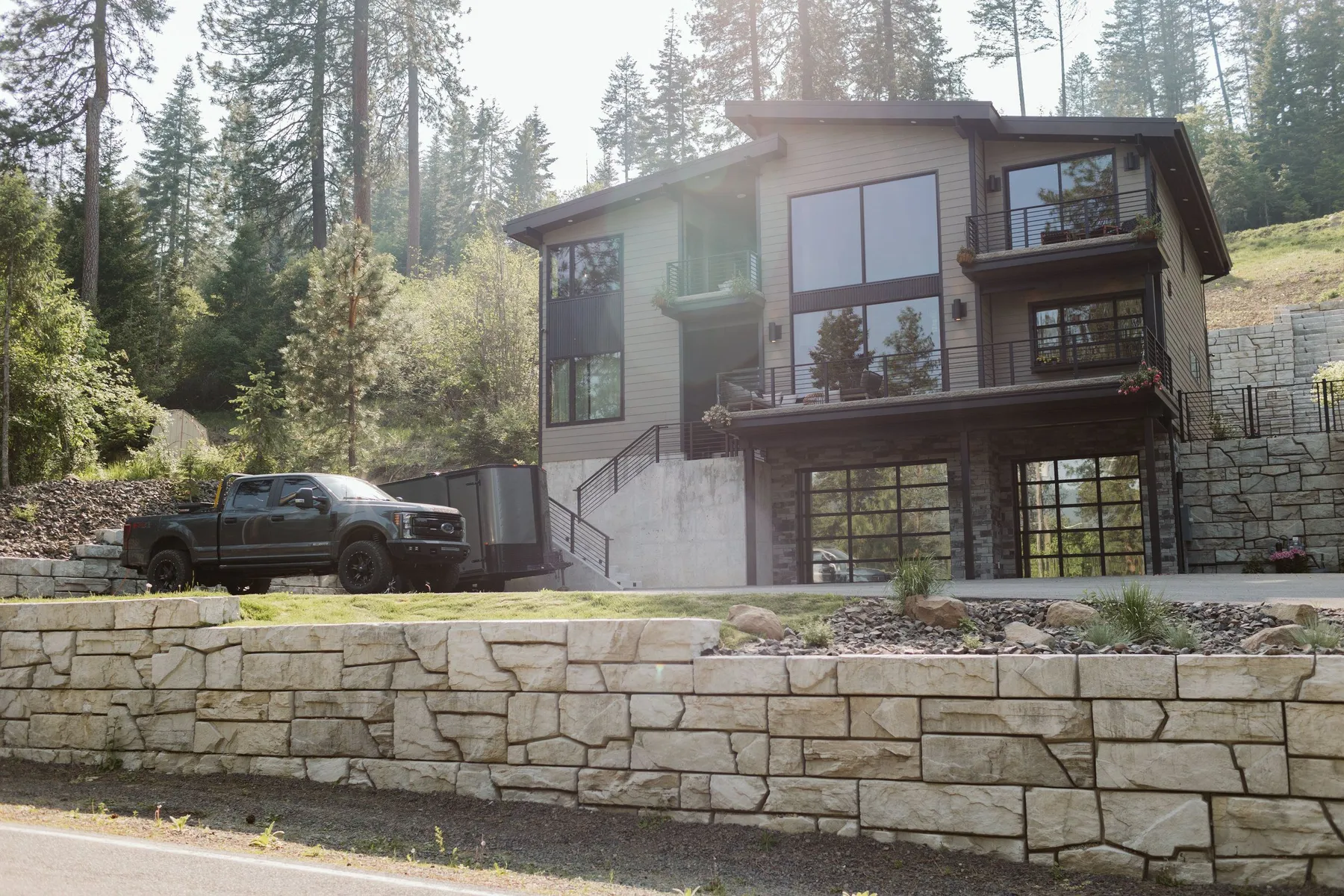 Lakeside home on a hillside above a long stone-textured block retaining wall along the road, with a pickup parked in front.