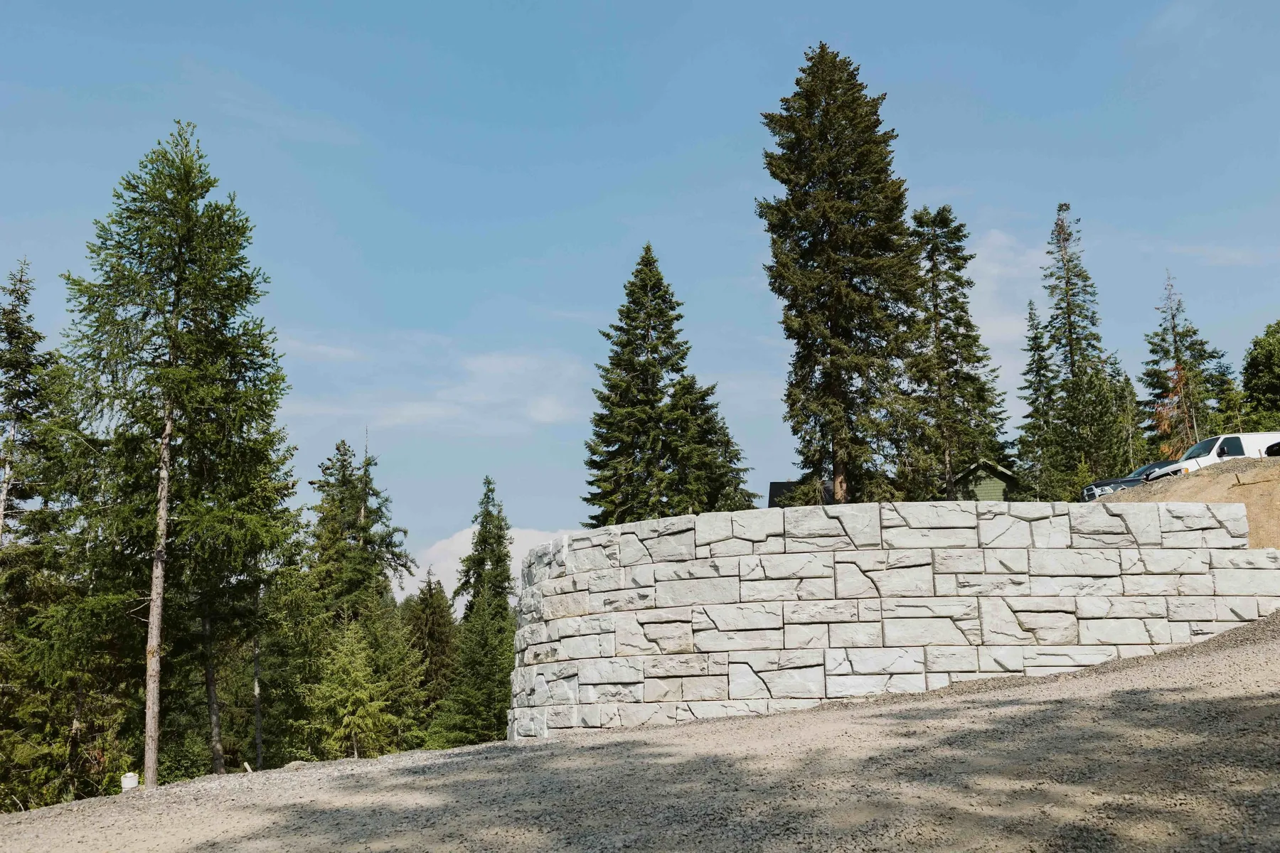 Curved retaining wall supporting the upper drive, with tall firs behind it under a blue sky.