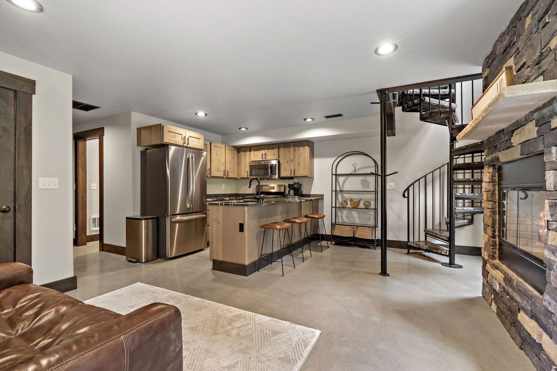 Living area with polished concrete floors, a black spiral iron staircase beside a stacked-stone fireplace and an open kitchen with bar seating.