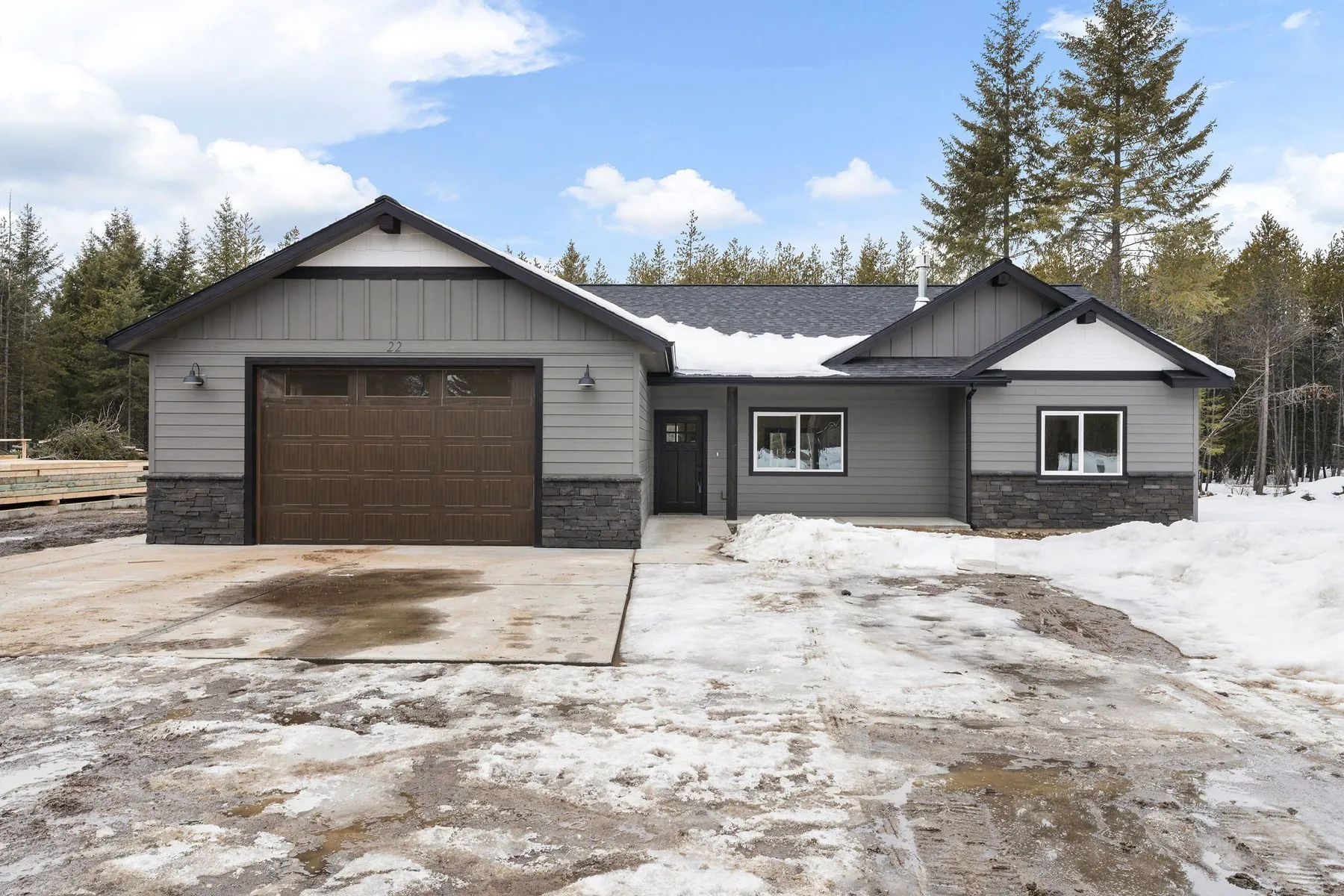 Front of the Timber & Treetops home, a gray single-story house with a brown garage door and stone base in a snowy forest clearing.