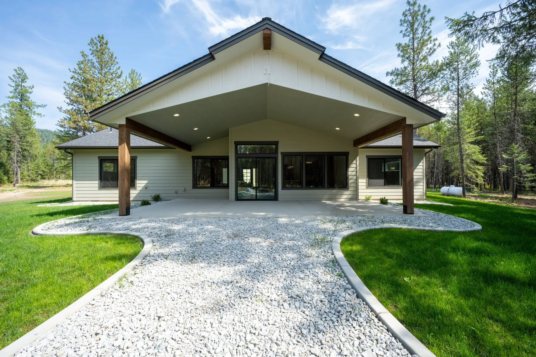 Blanchard Bliss, a light gray ranch home with a deep covered patio on timber posts, a curving white-rock path and green lawn among pines.