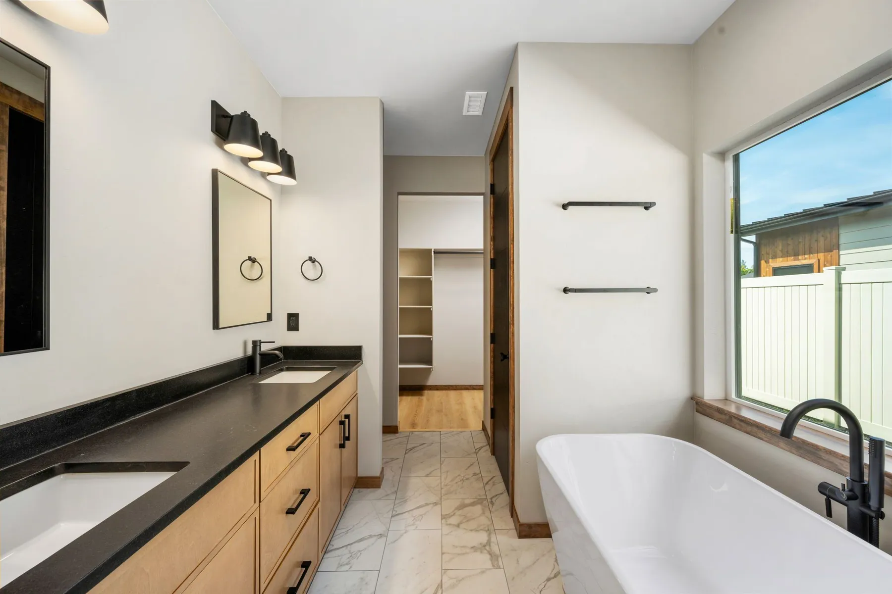 Primary bathroom with a black-topped dual vanity, a freestanding soaking tub under a window and the walk-in closet beyond.