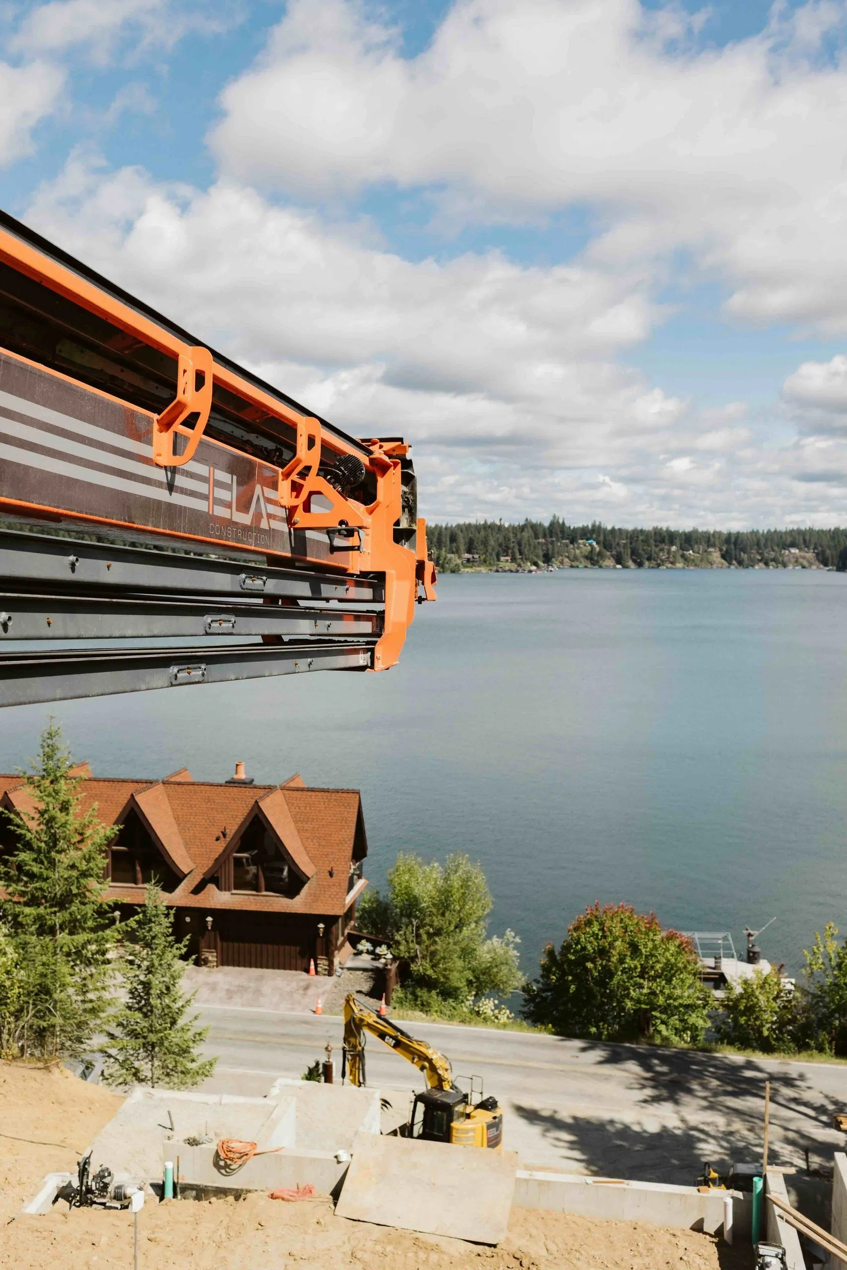 Boom of an orange drill rig over the hillside site, with the lake and a lakeside home below.