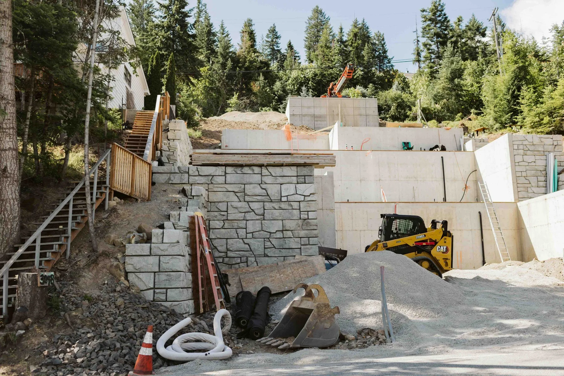 Stepped concrete and stone-block walls on the house pad, with a skid steer, gravel pile and wooden stairs up the slope.