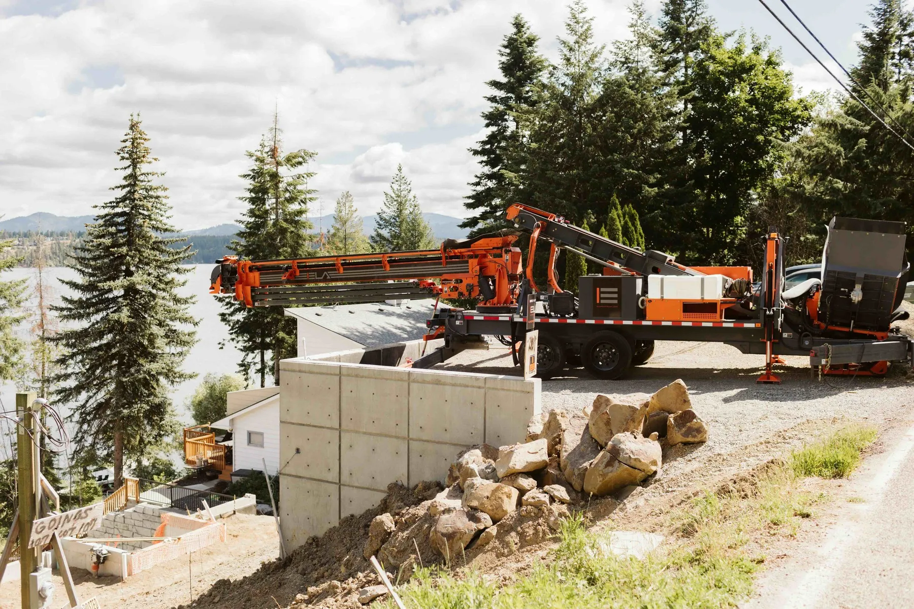 Orange anchor drill rig set up on a concrete wall above the lake, with boulders stacked on the slope.