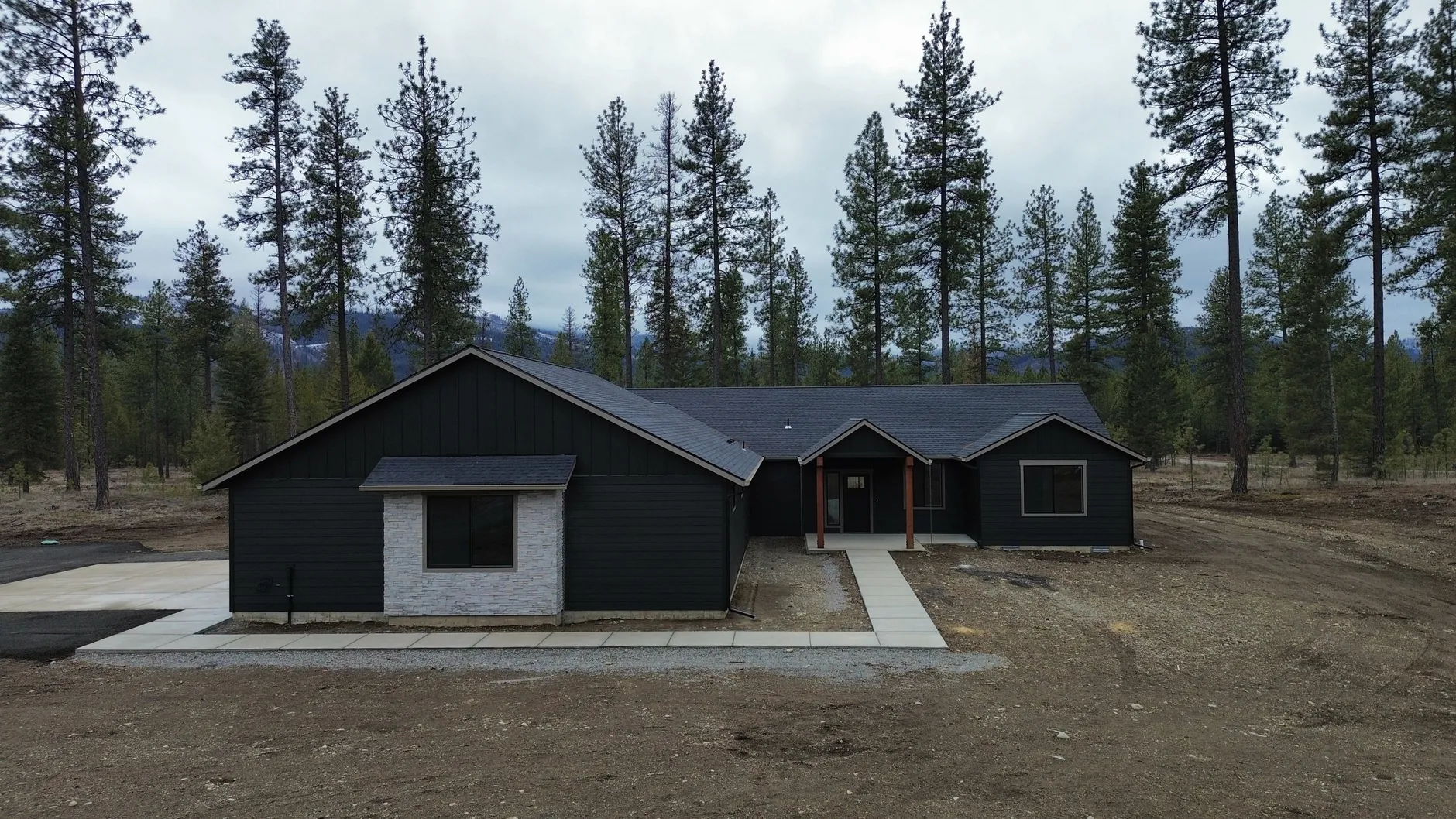 Front of The Summit, a dark single-story home with a white stone accent and a covered timber entry on a newly graded lot among pines.