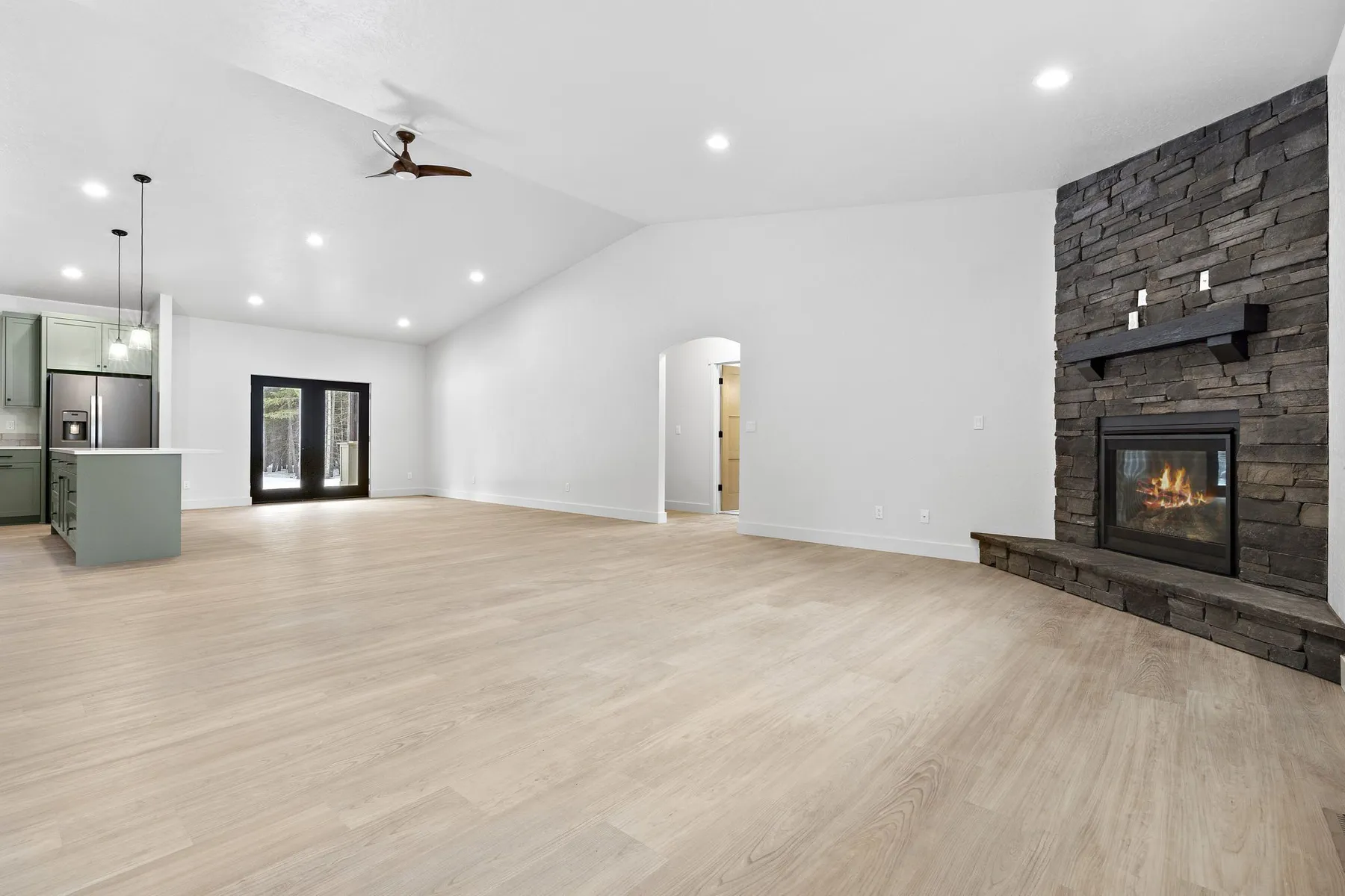 Empty great room with light plank flooring, an arched doorway and a corner stone fireplace with a dark wood mantel.