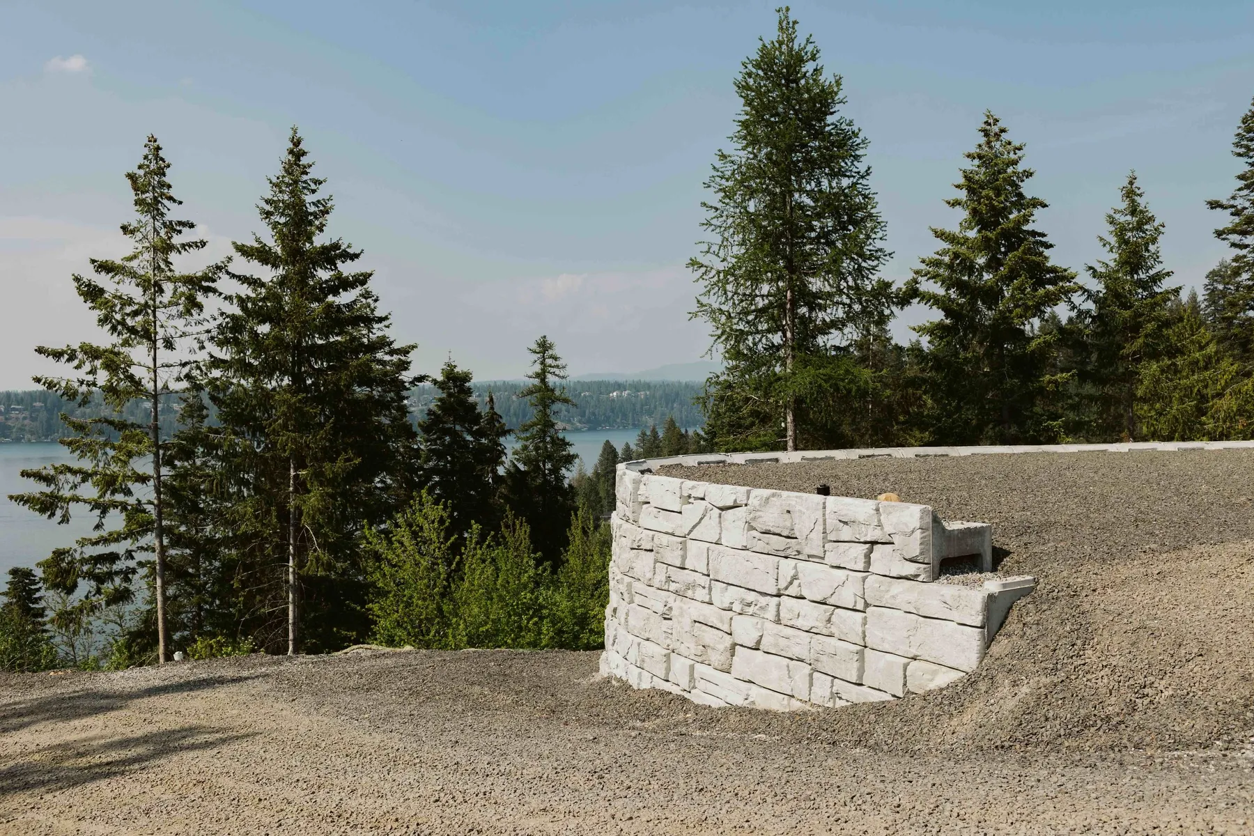 Curved block retaining wall at the edge of the graded pad, with evergreens and the lake beyond.
