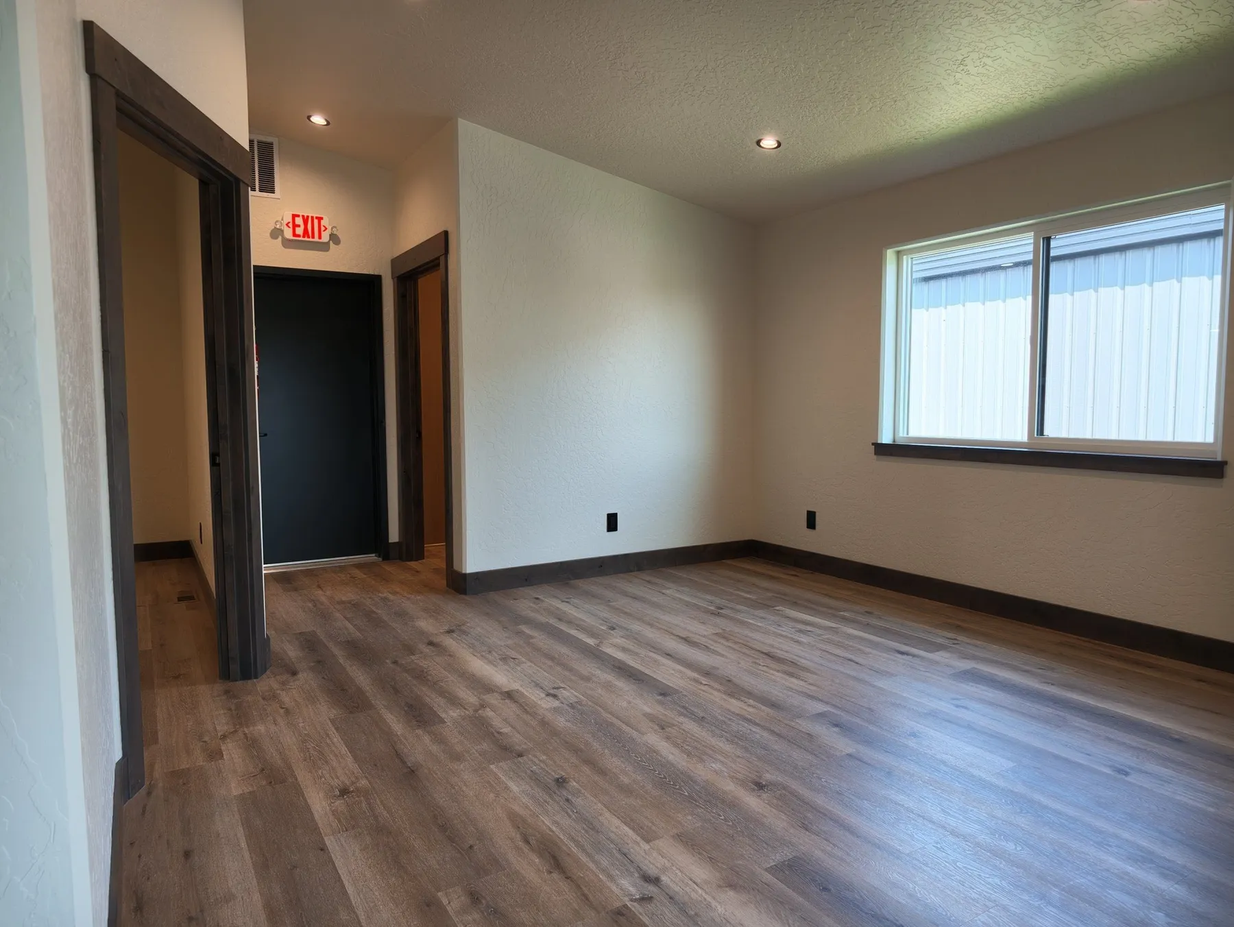 Open office space with wood-look flooring, a lit exit sign over a black door and windows along one wall.