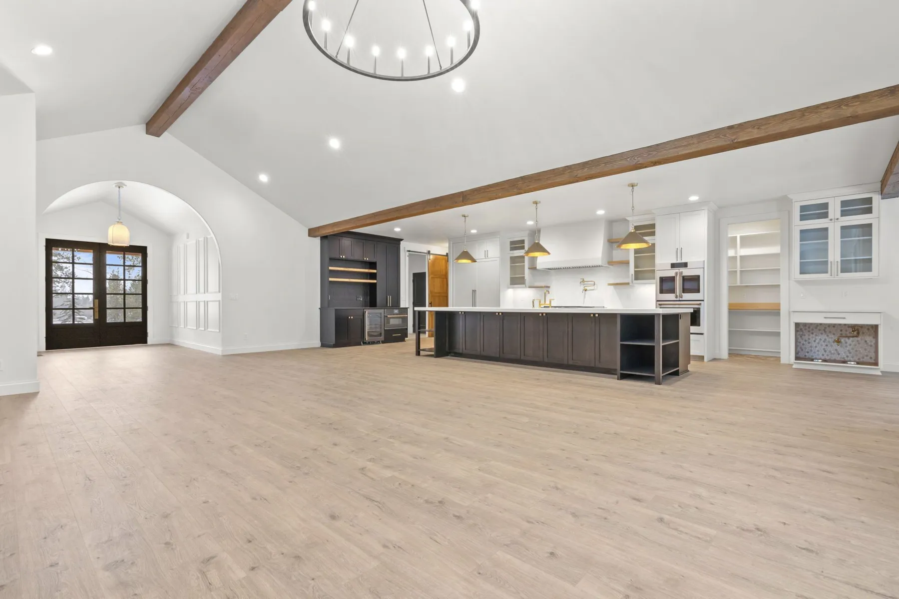 Open great room looking toward the kitchen, with a long dark island, a cedar ceiling beam and an arched doorway to the side.
