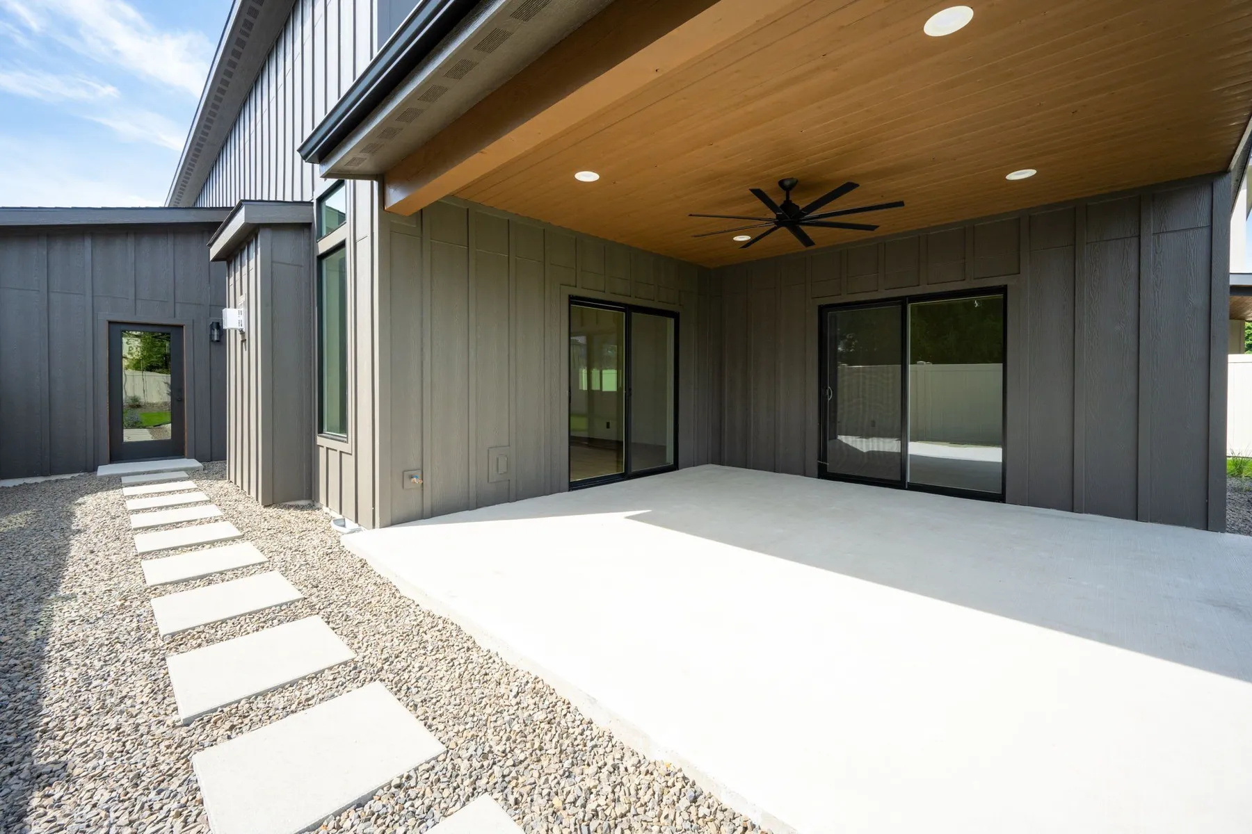 Covered back patio with a wood-plank ceiling and fan, sliding doors and stepping stones across a gravel side yard.
