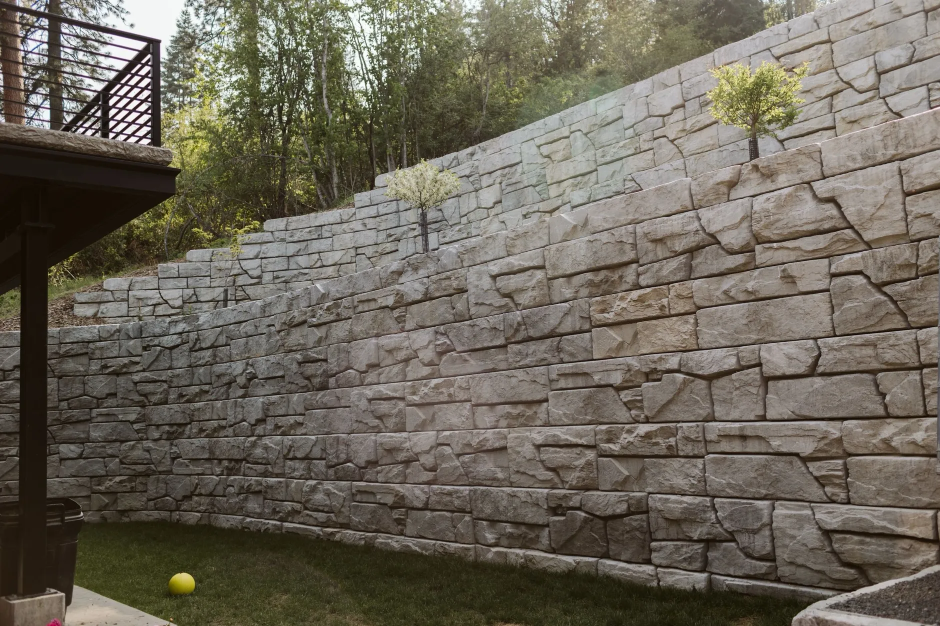 A multi-tiered stone-textured retaining wall with small trees planted in its terraces, rising above a lawn beside a covered deck in the Hayden Lake hills.