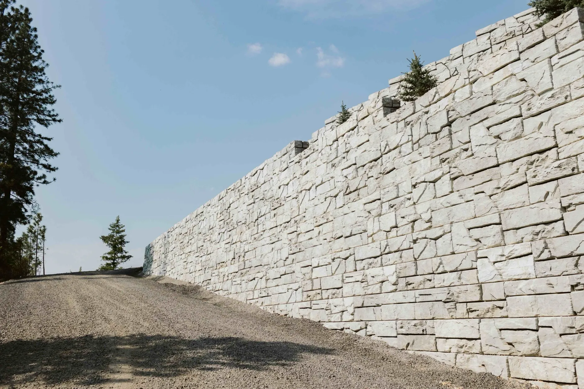 A tall stone-textured block retaining wall running alongside a gravel driveway that climbs a hillside above Hayden Lake.