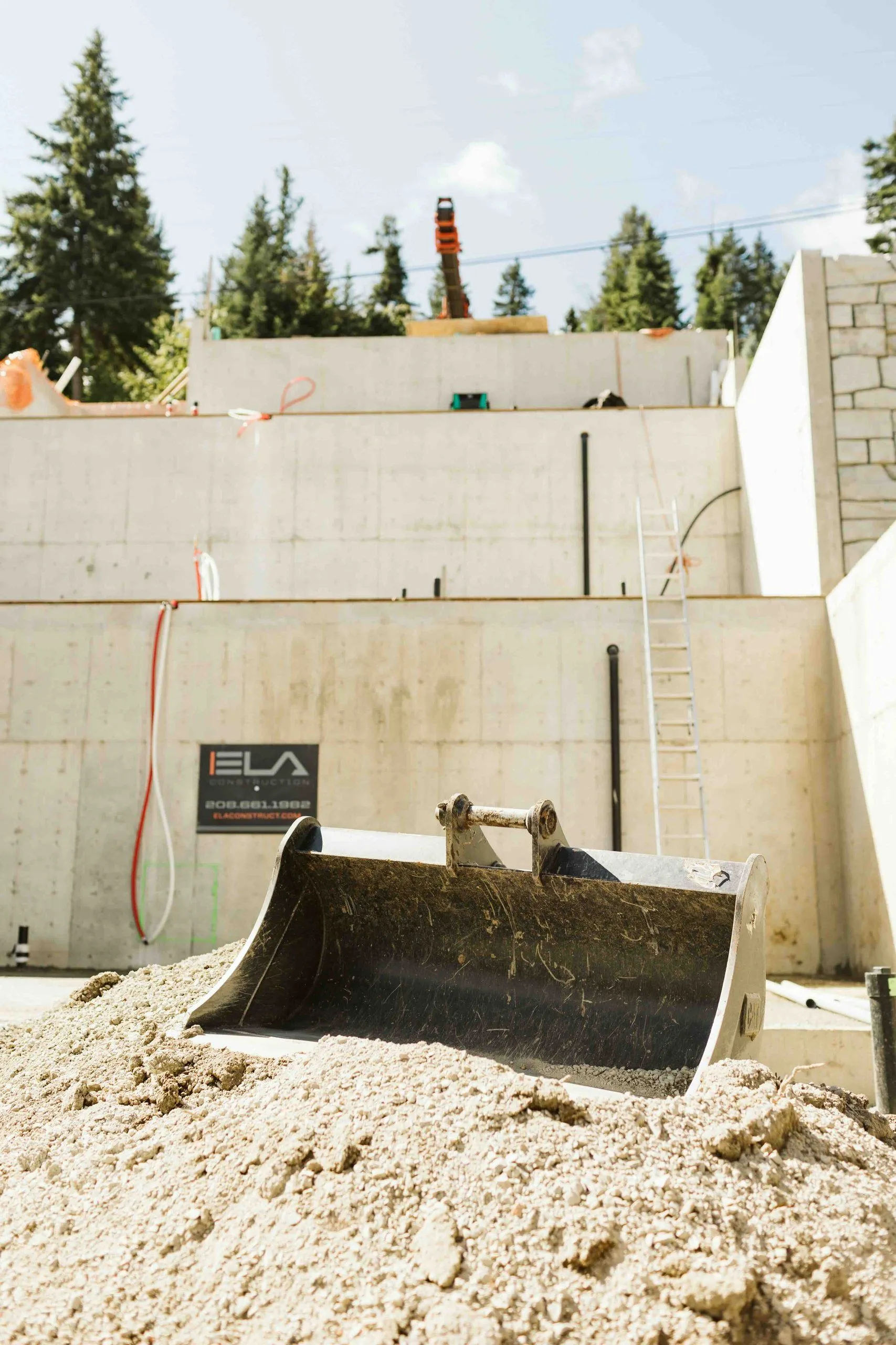 Excavator bucket resting on gravel in front of stepped concrete walls marked with an Ela Construction sign.