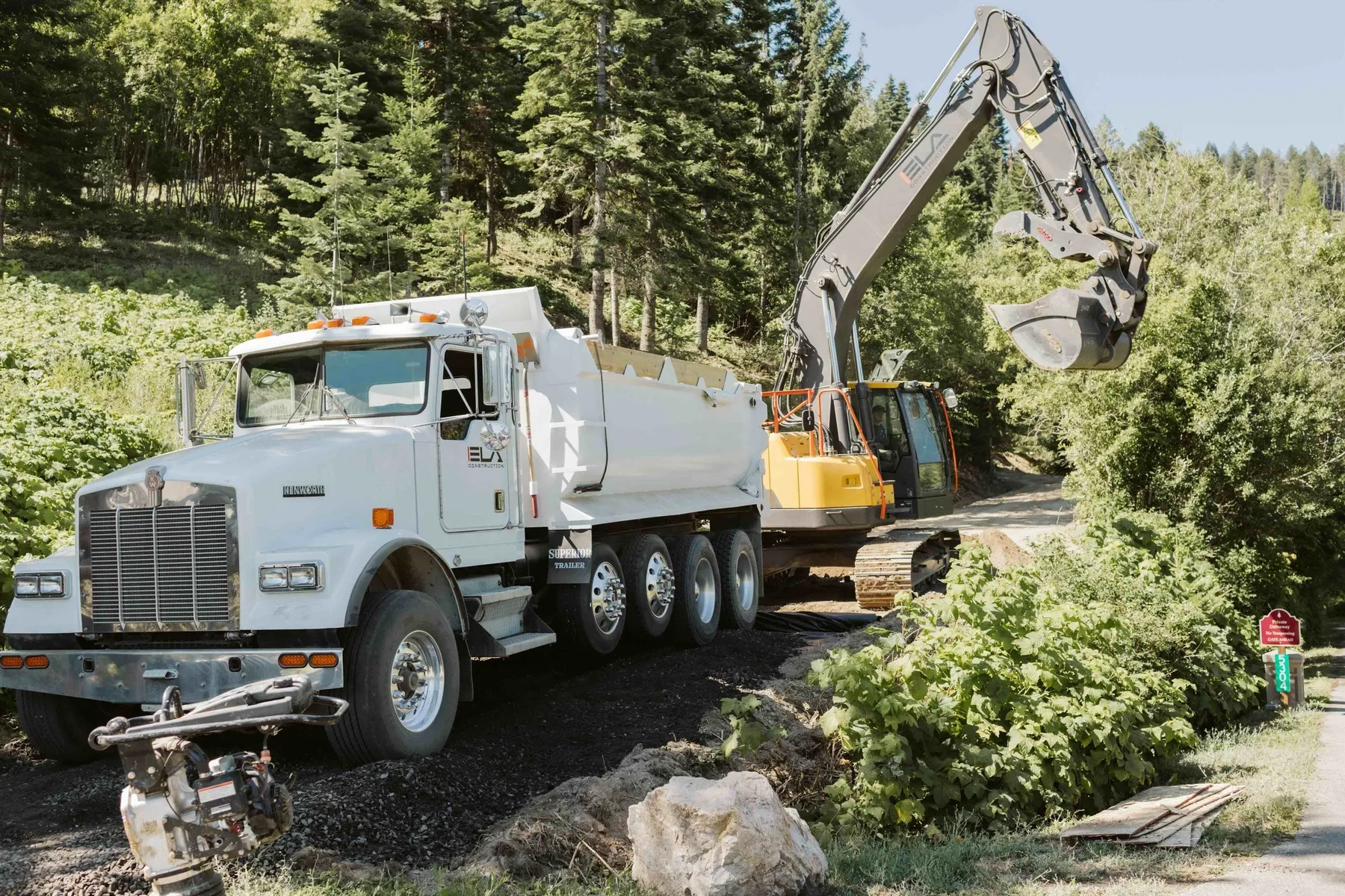 Excavator loading gravel from a white dump truck for the driveway base layer, with a plate compactor in front.