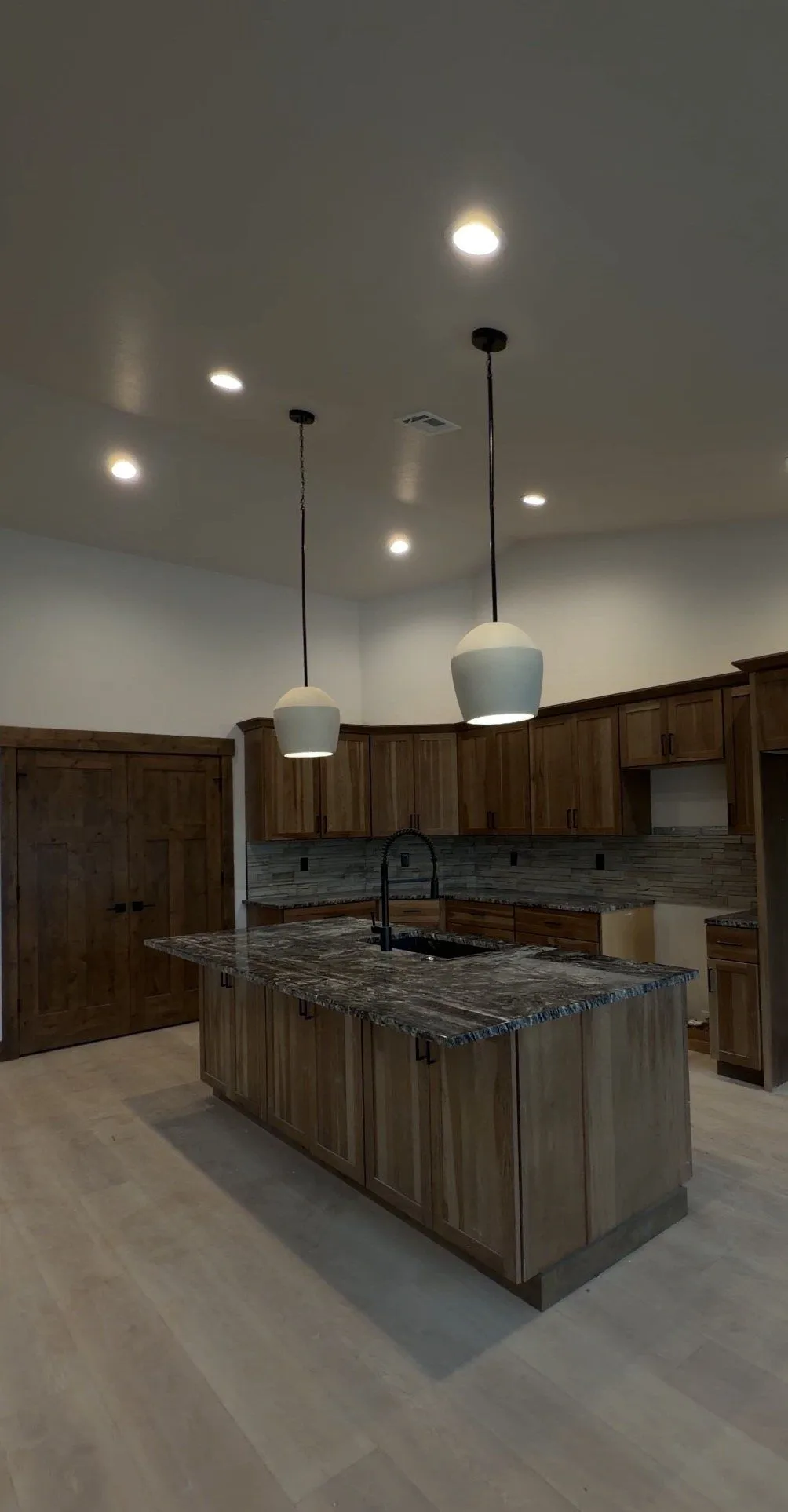 Kitchen with rich wood cabinets, a dark granite island, two white pendant lights and light plank flooring.