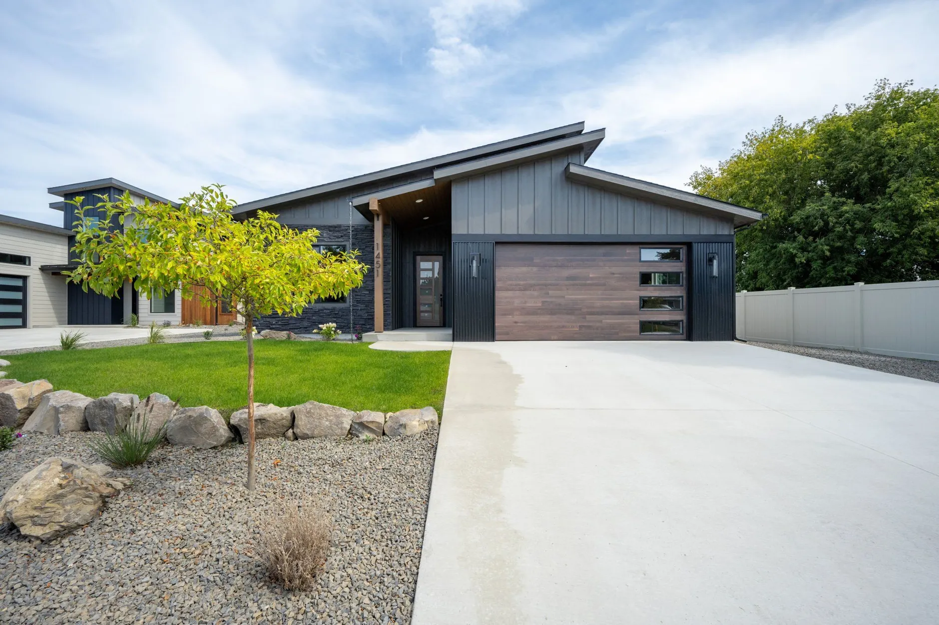 Modern single-story home with dark board-and-batten siding, a shed roof, a wood-look garage door and rock landscaping along the driveway.