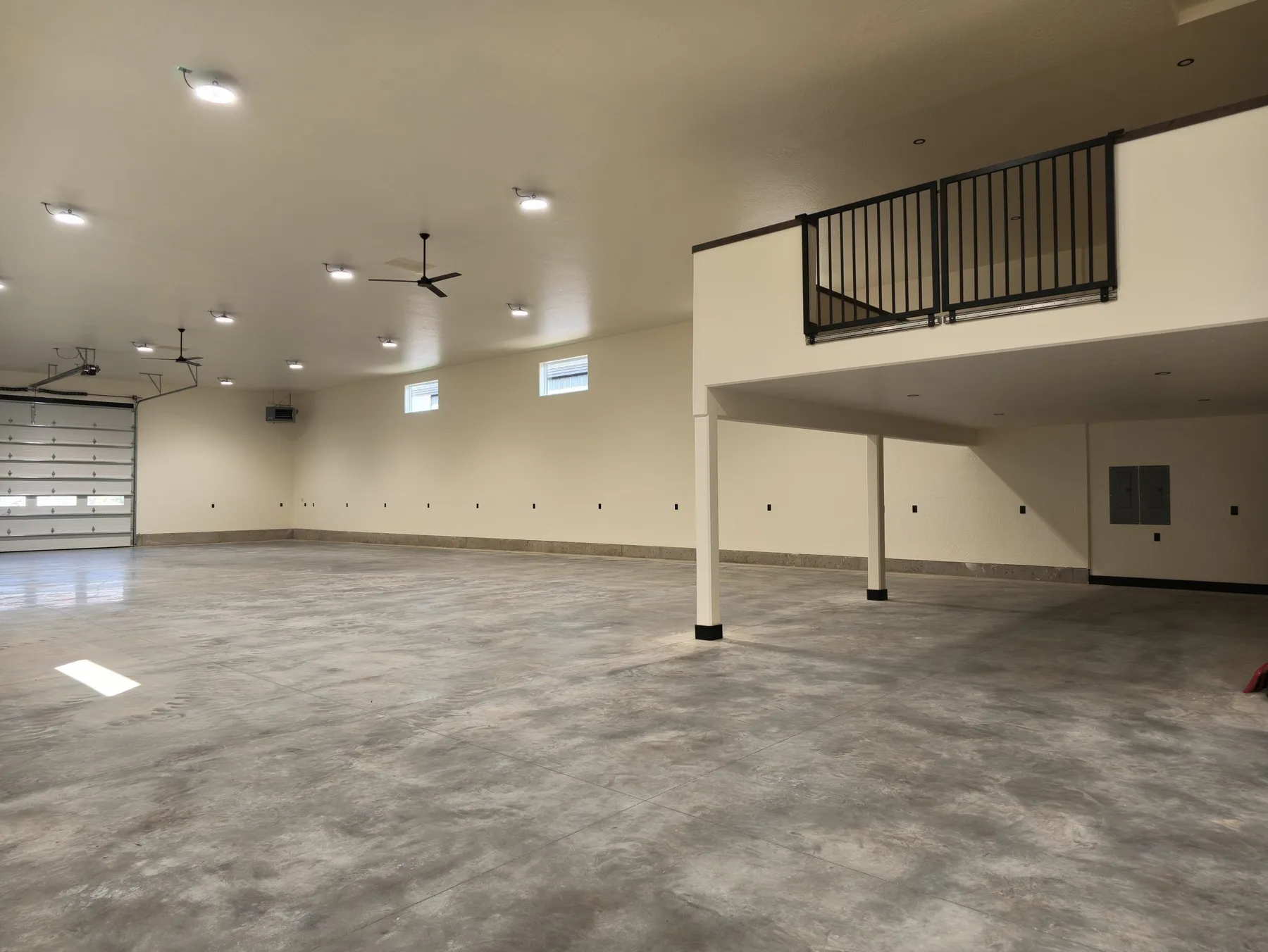 Warehouse interior with an overhead door, ceiling fans and an upper mezzanine office with a black metal railing.