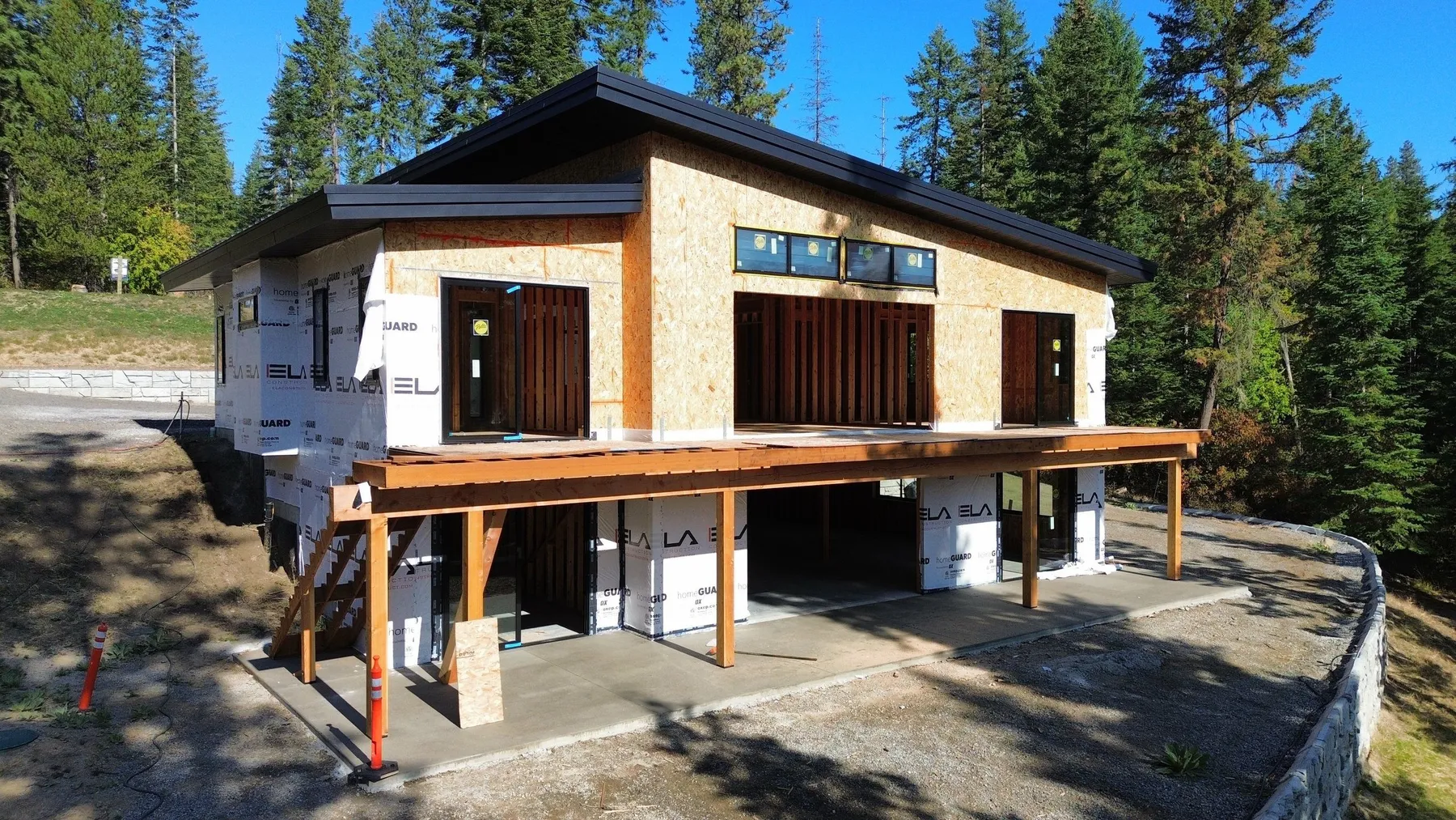 Lake-side elevation of the house under construction with exposed sheathing, large window openings and a timber-framed deck above the patio.