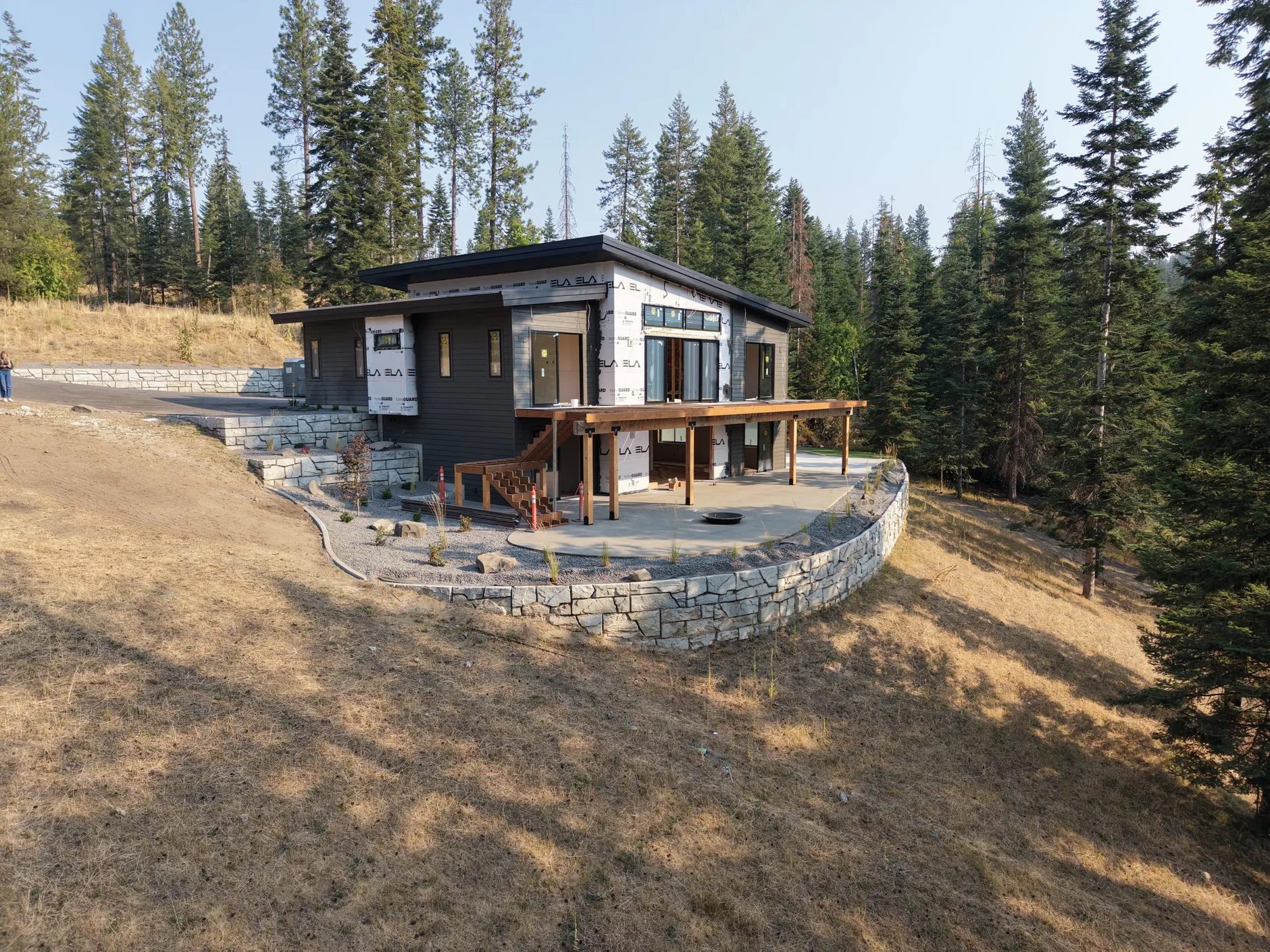 Two-story modern home under construction above Hayden Lake, wrapped in house wrap, with a timber deck and a curved stone retaining wall on a dry hillside.