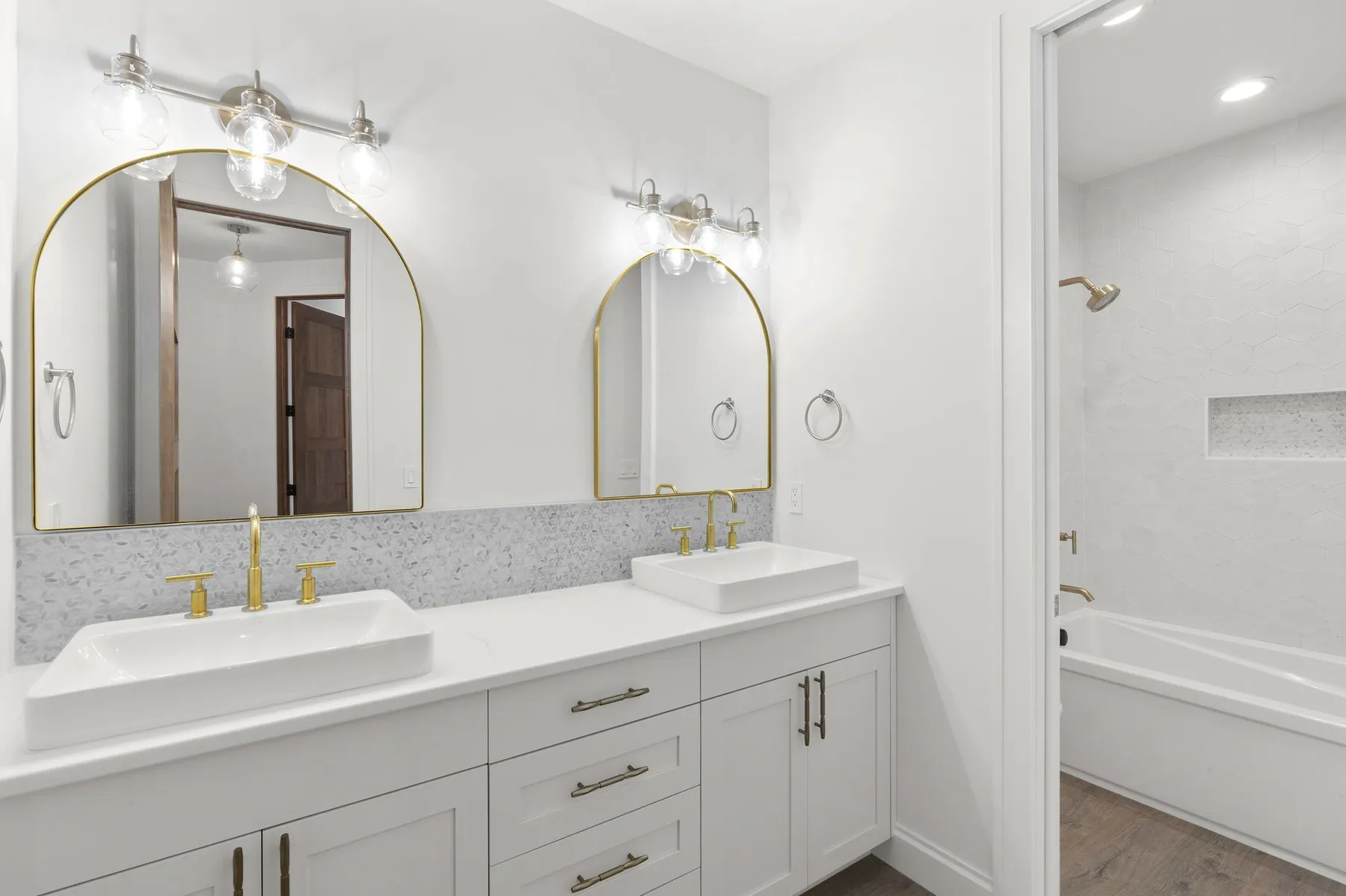 Bathroom with a white double vanity, arched brass mirrors and a tub-shower combination through the doorway.