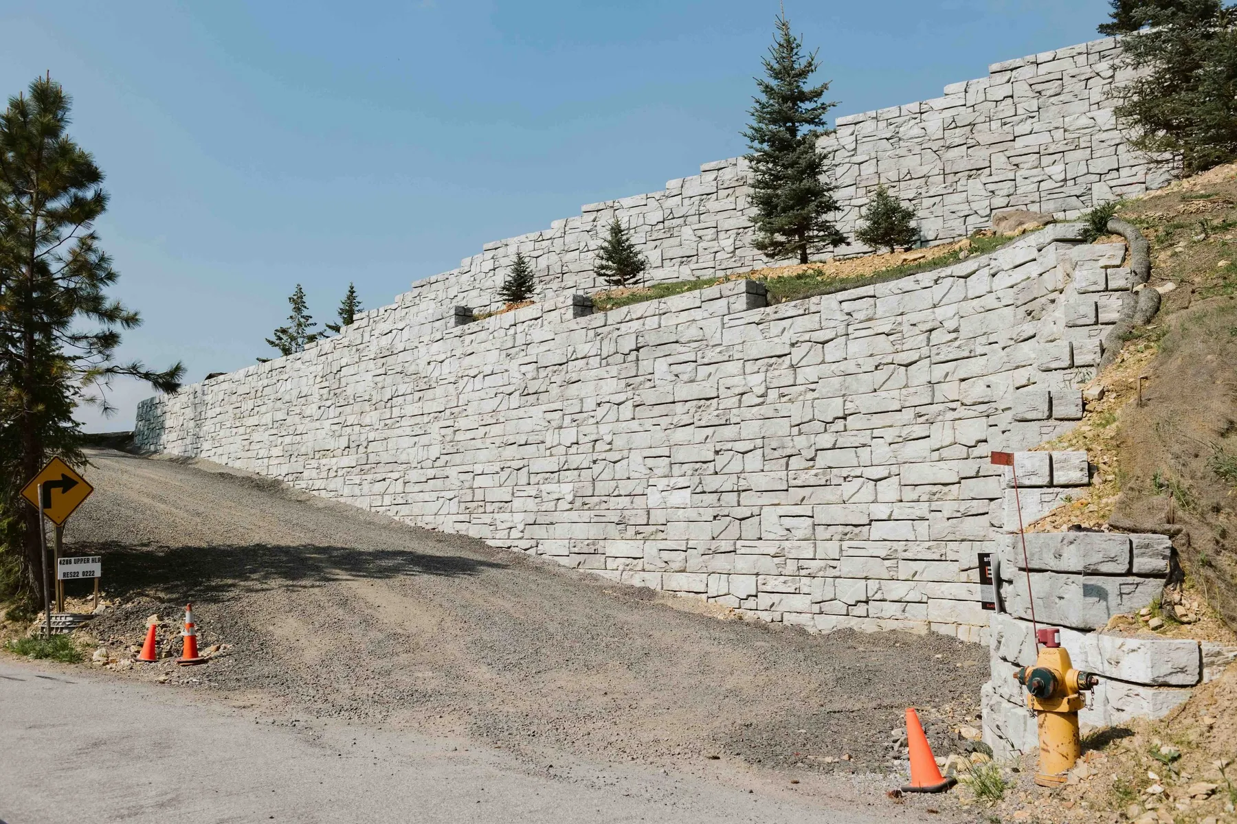 Massive stone-textured block retaining wall in two tiers along a steep driveway, with traffic cones at its base.