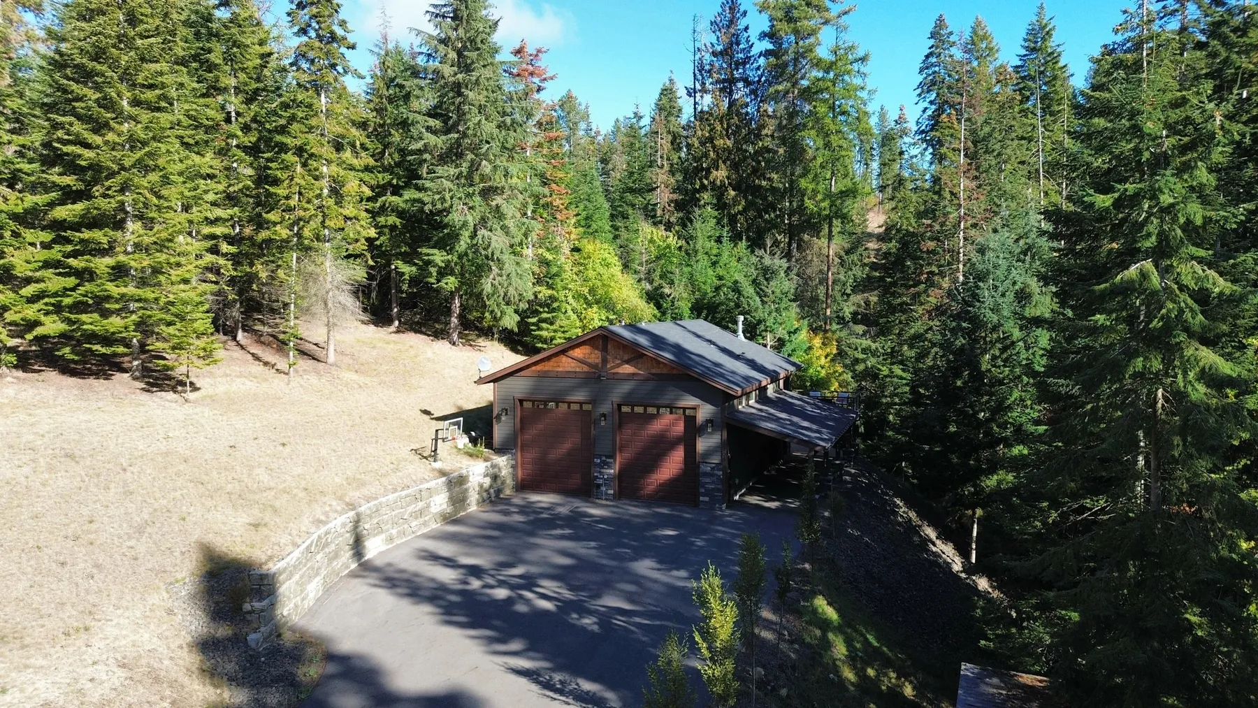 Aerial view of the detached two-bay shop with an ADU, a paved driveway and a stone retaining wall in a forested clearing.