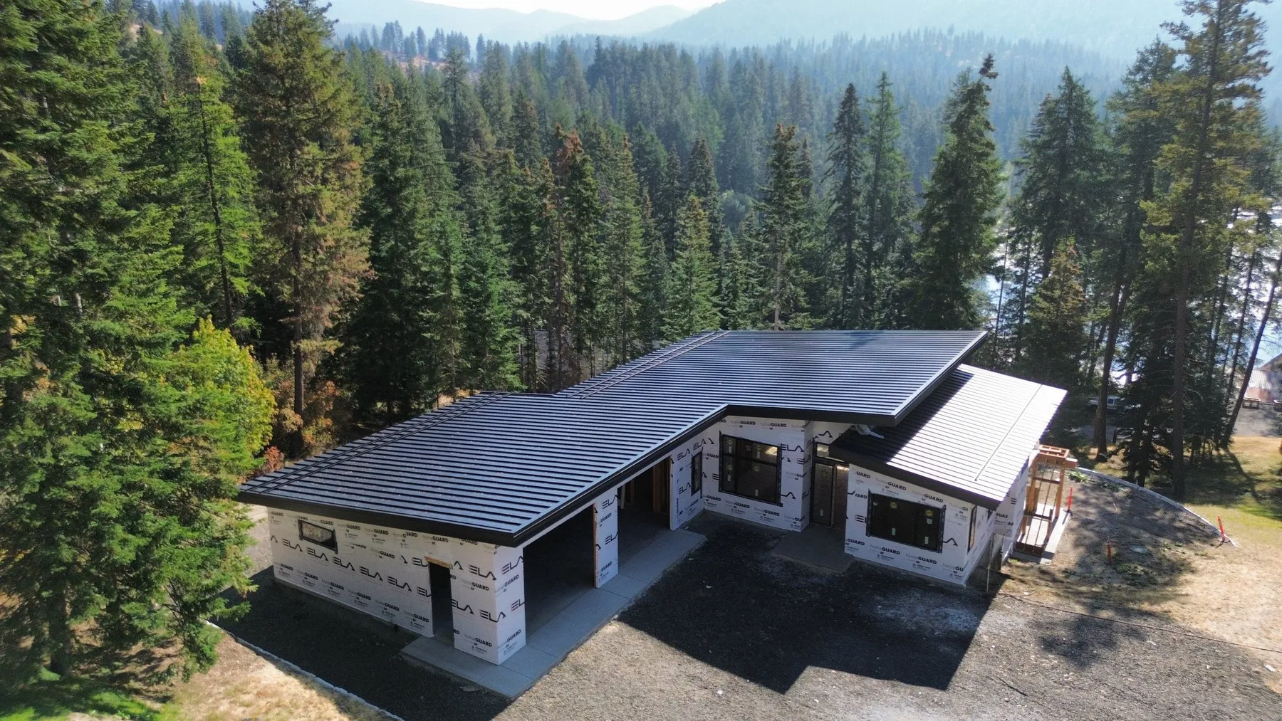 Aerial view of the home under construction, its black metal roof and house-wrapped walls in a clearing of tall evergreens.