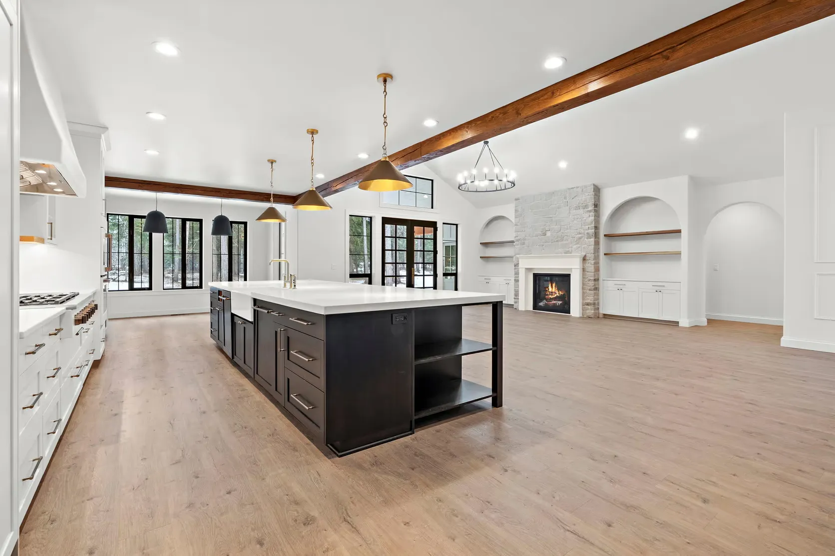 An open floor plan interior of a custom home with wide-plank floors, a gas fireplace, exposed wood beams, white quartz countertops, a farmhouse sink, and champagne bronze and matte black finishes.