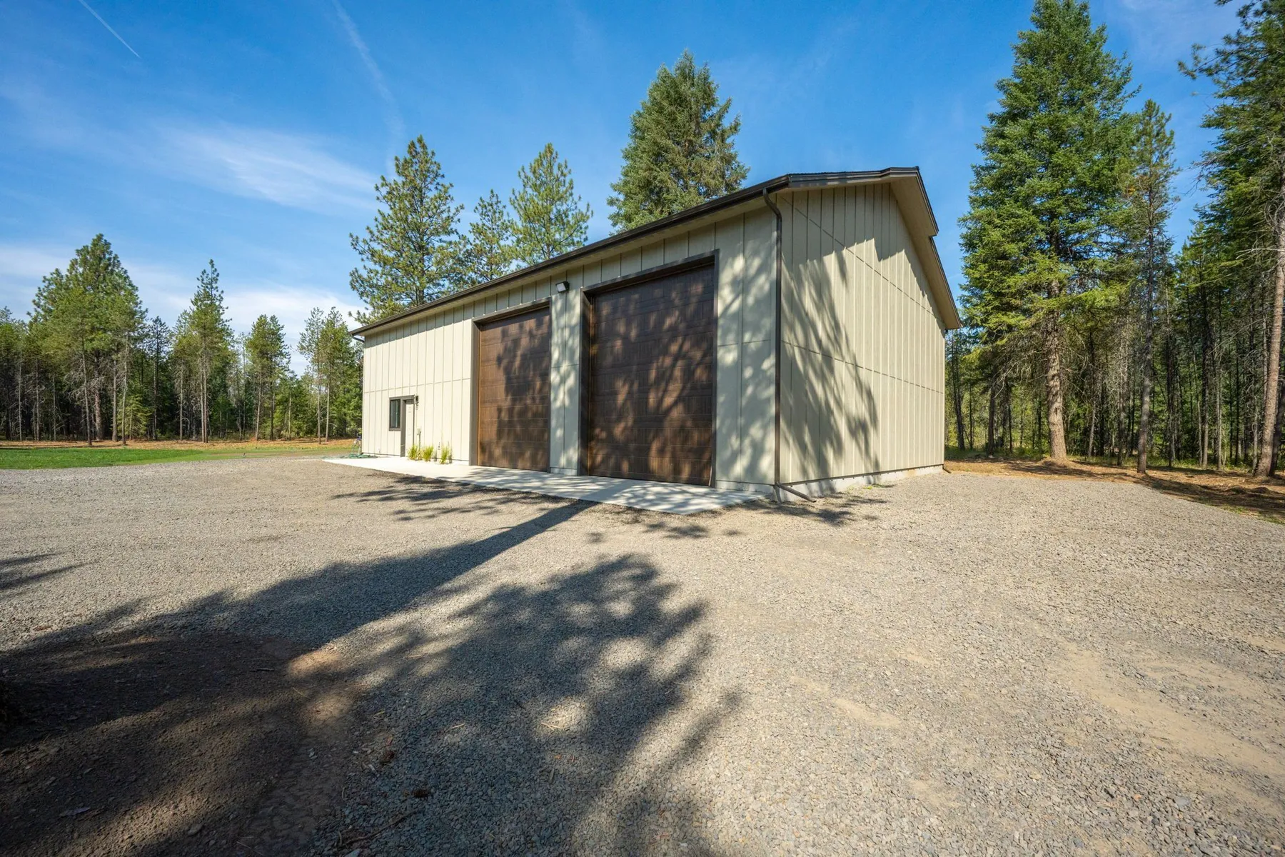 Detached custom shop with two tall brown overhead doors on a gravel pad among pines.