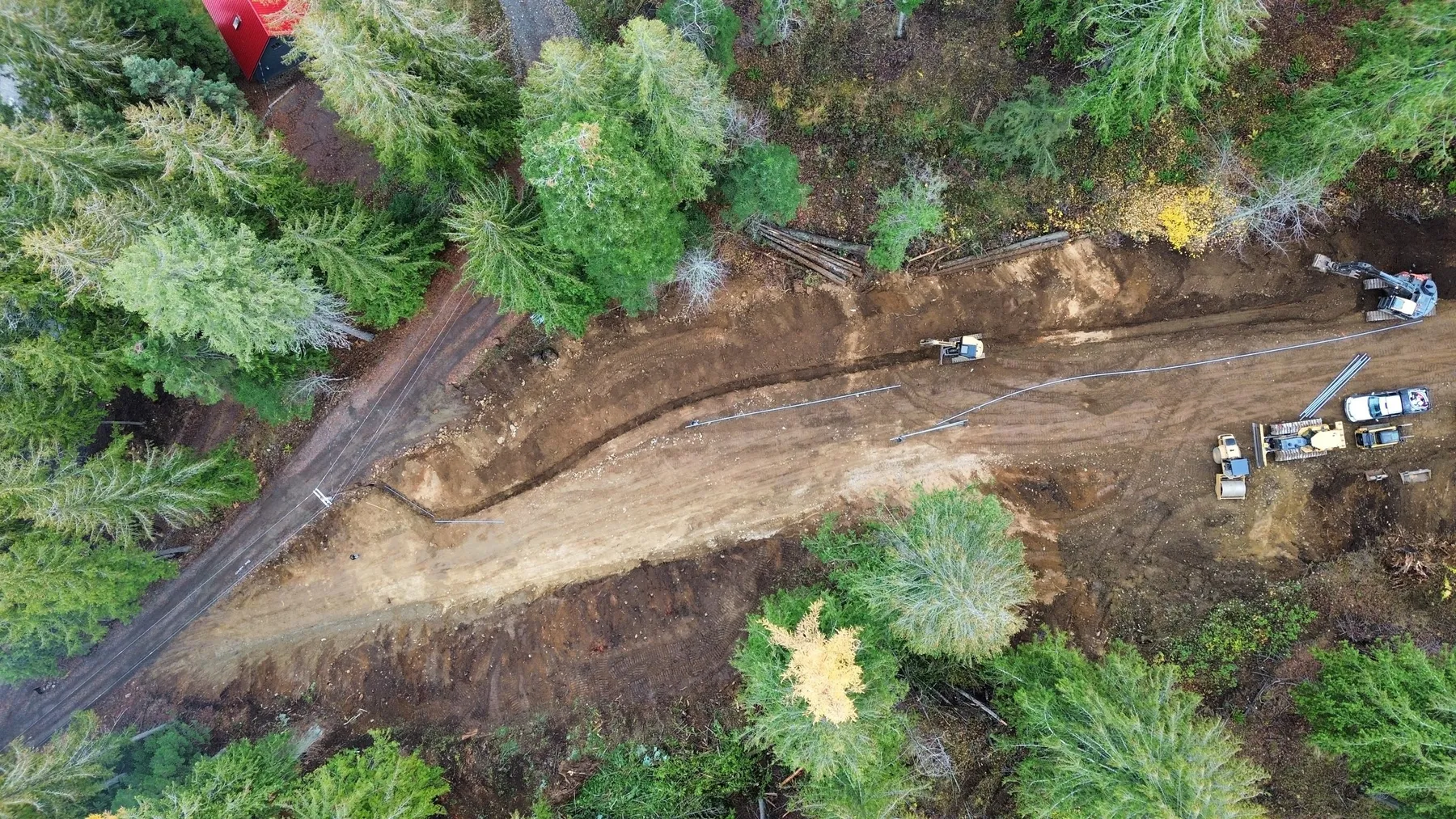 Aerial view of the new road cut through the forest, with conduit laid in a trench alongside it and an excavator and trucks at work.