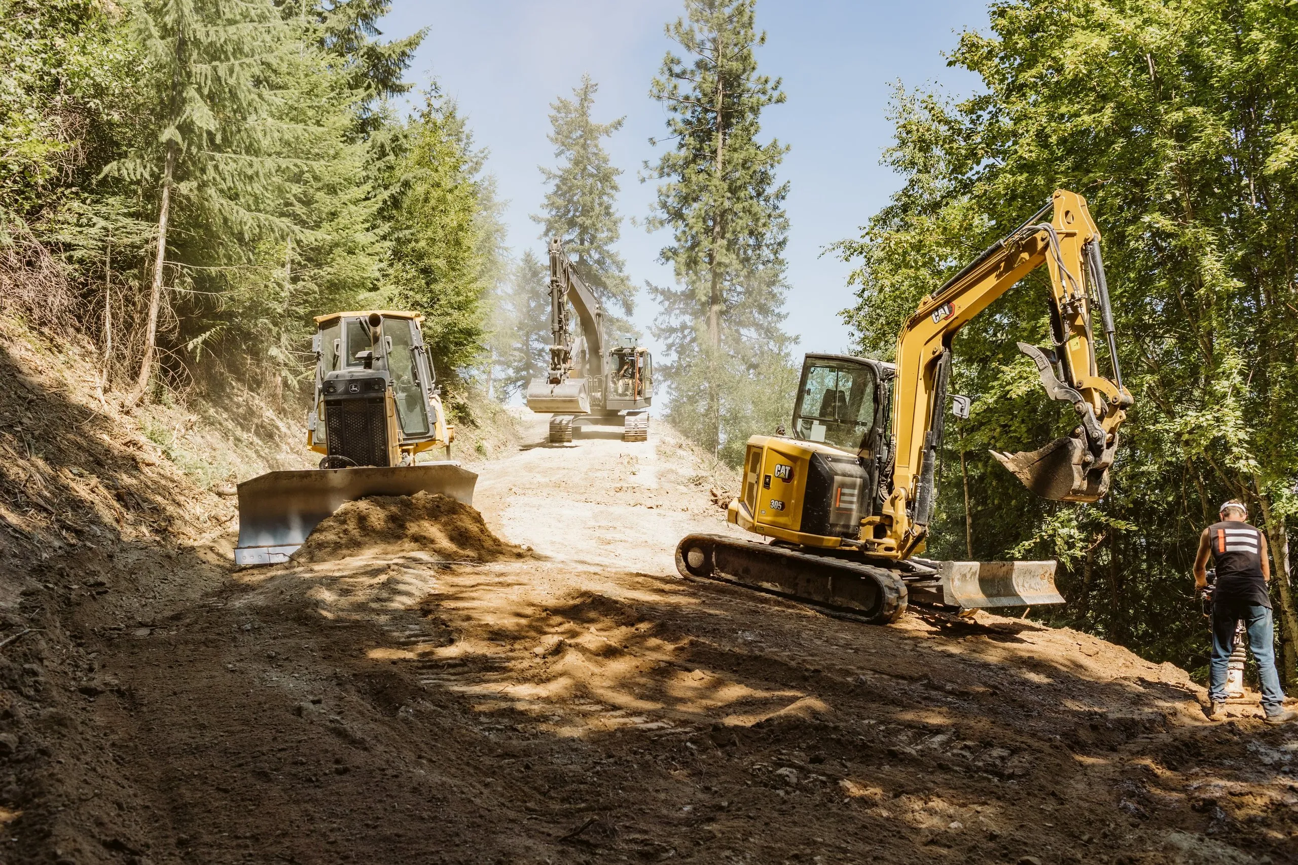 Two yellow excavators and a dozer cutting a dirt access road up a forested hillside, with an Ela crew member in a hi-vis shirt watching from the right.