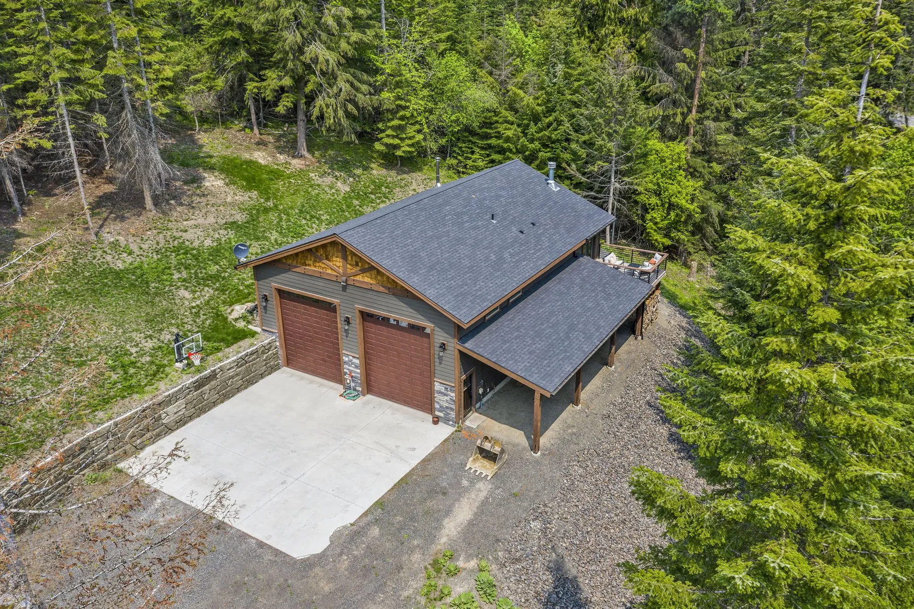 Aerial view of the shop home, a gray two-bay shop with wood garage doors, a covered side porch and a concrete apron in a forested clearing.