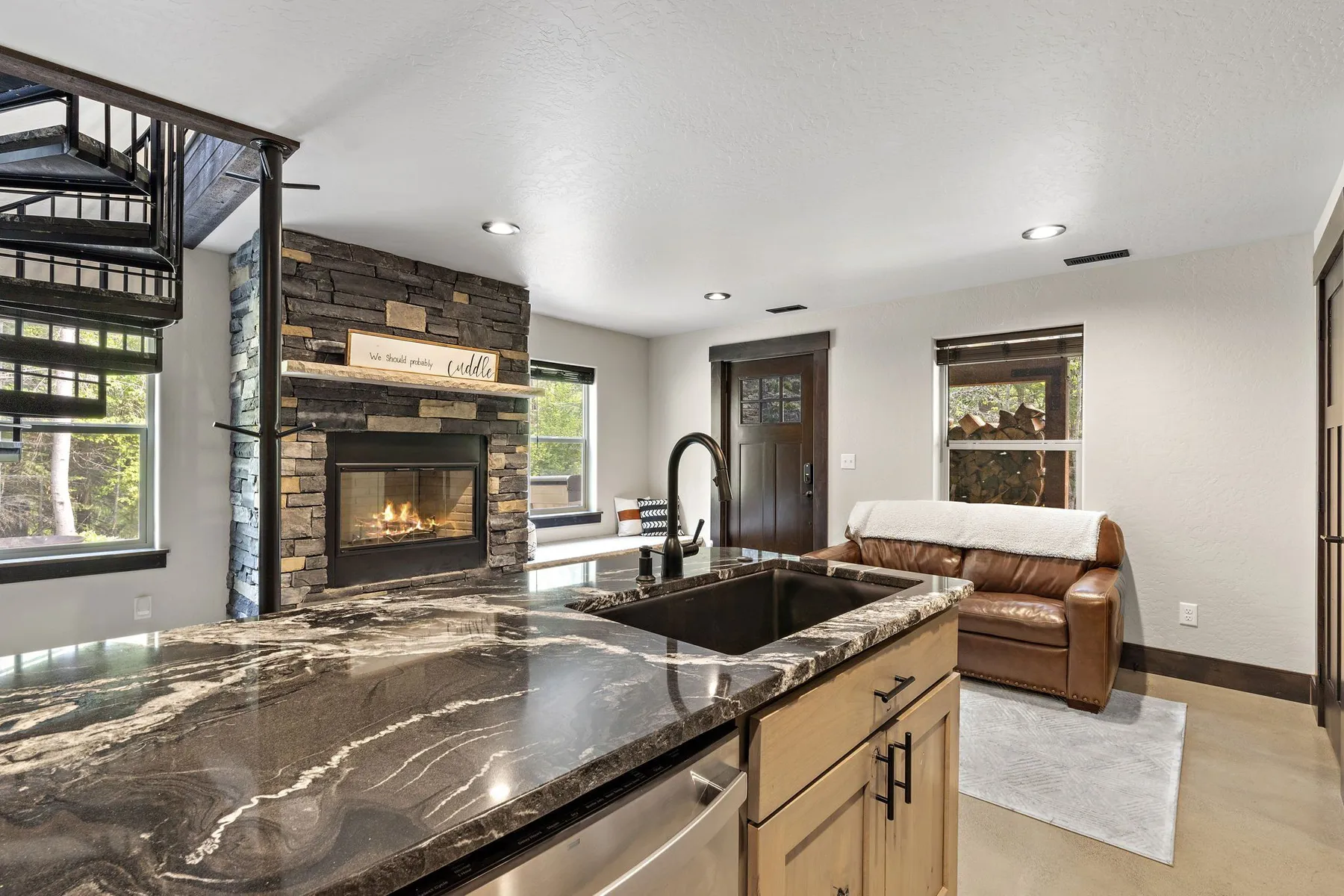 Kitchen island with a dark marbled stone countertop and black sink, facing the stone fireplace and a leather sofa.
