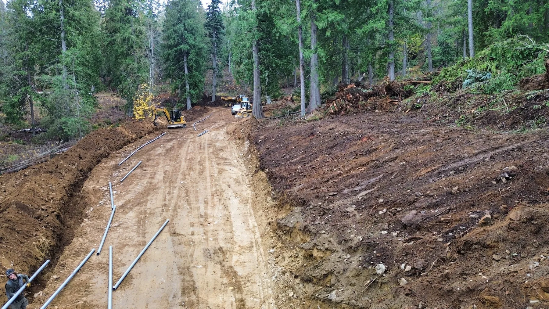 Utility trench running beside the new gravel road, with gray conduit pipe and an excavator working farther up the grade.