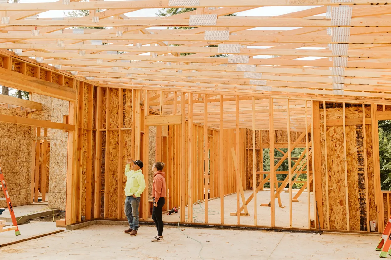 Two people inspecting wooden framing inside a house under construction with exposed ceiling beams and wall studs.