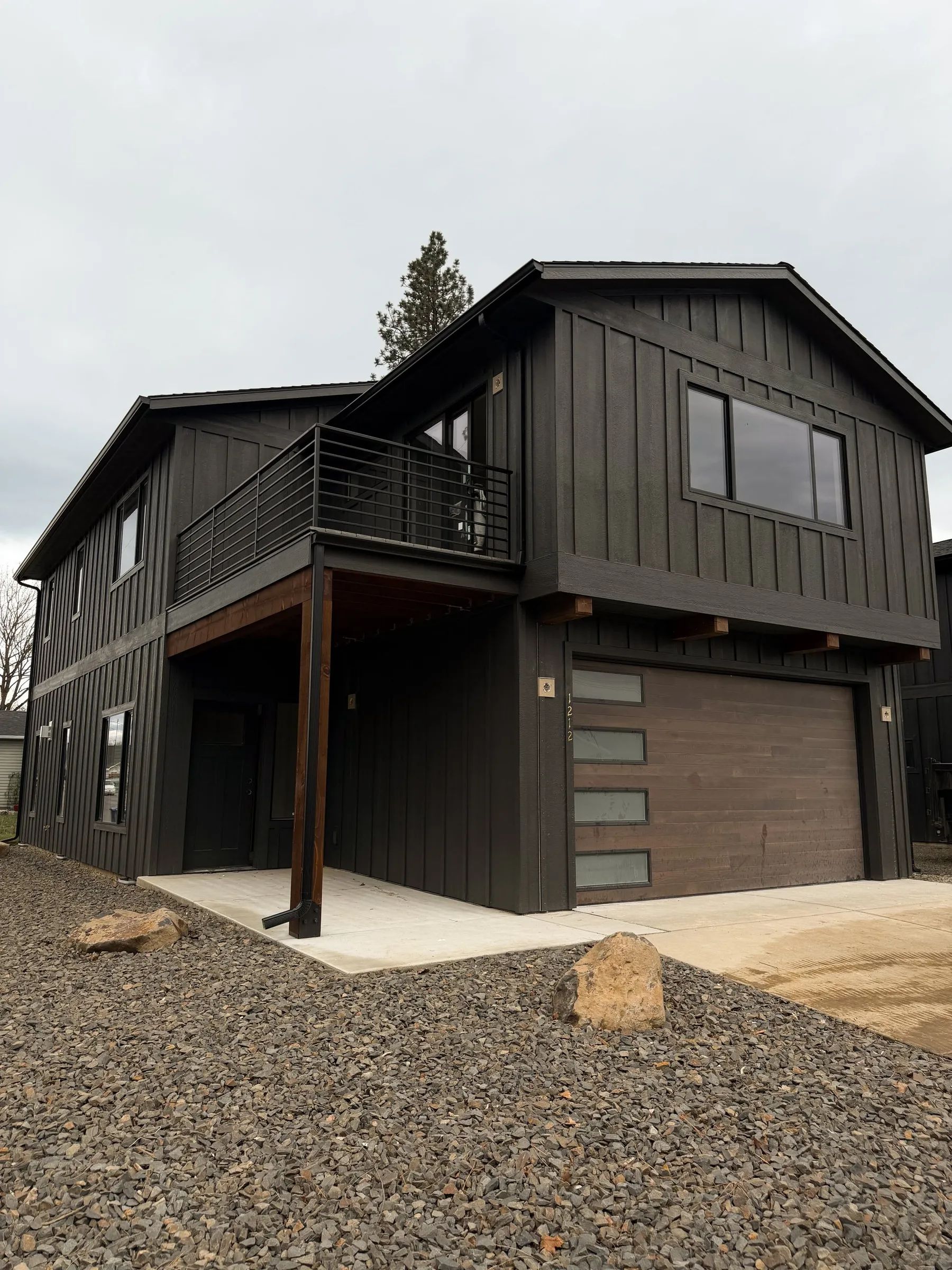 Finished two-story townhome in dark board-and-batten siding with a covered entry, black balcony railing and wood-look garage door above a gravel yard.