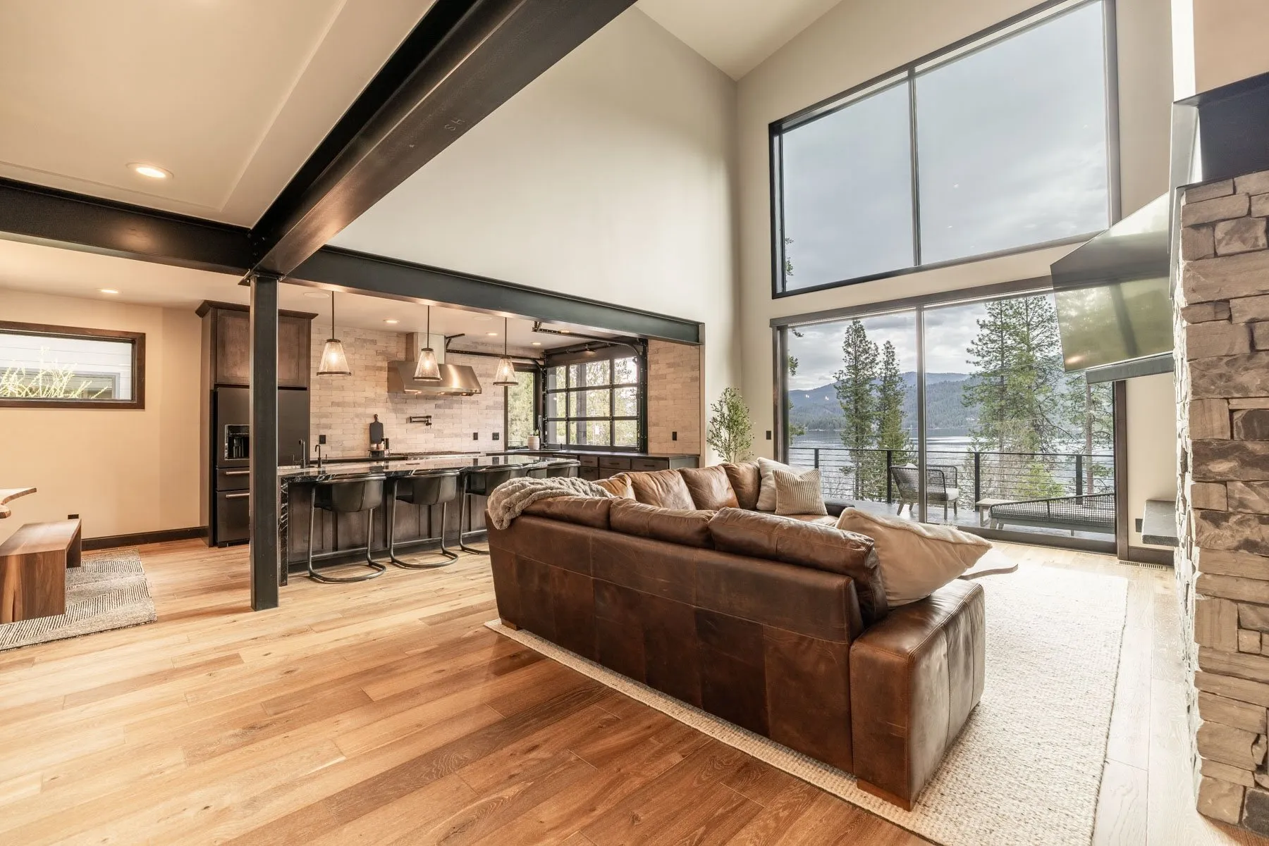 Great room with exposed black steel beams, a leather sectional and tall windows framing the lake, open to the kitchen behind.