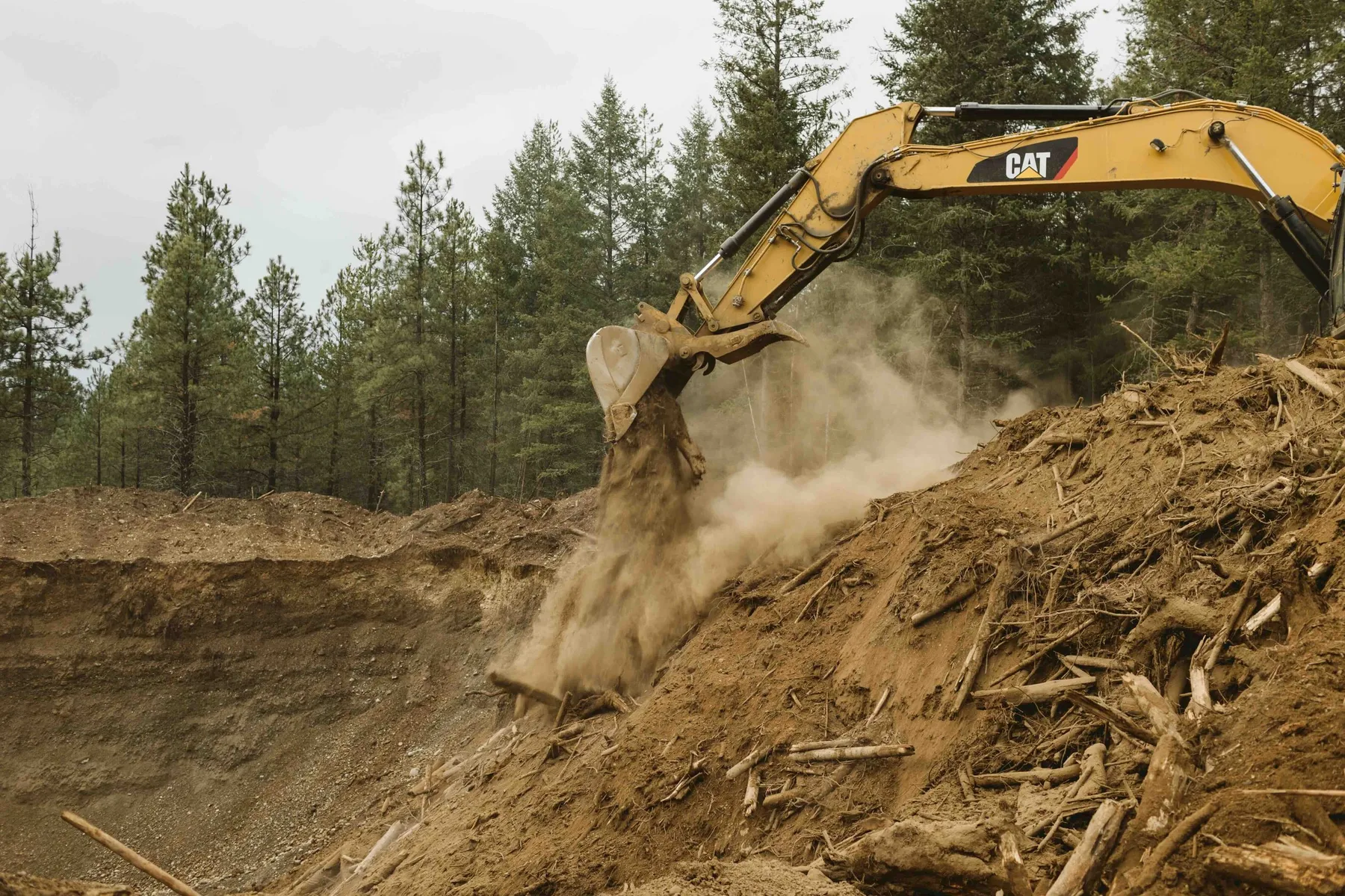CAT excavator digging into a pile of roots and soil while clearing land at Clagstone Estates, with dust in the air.
