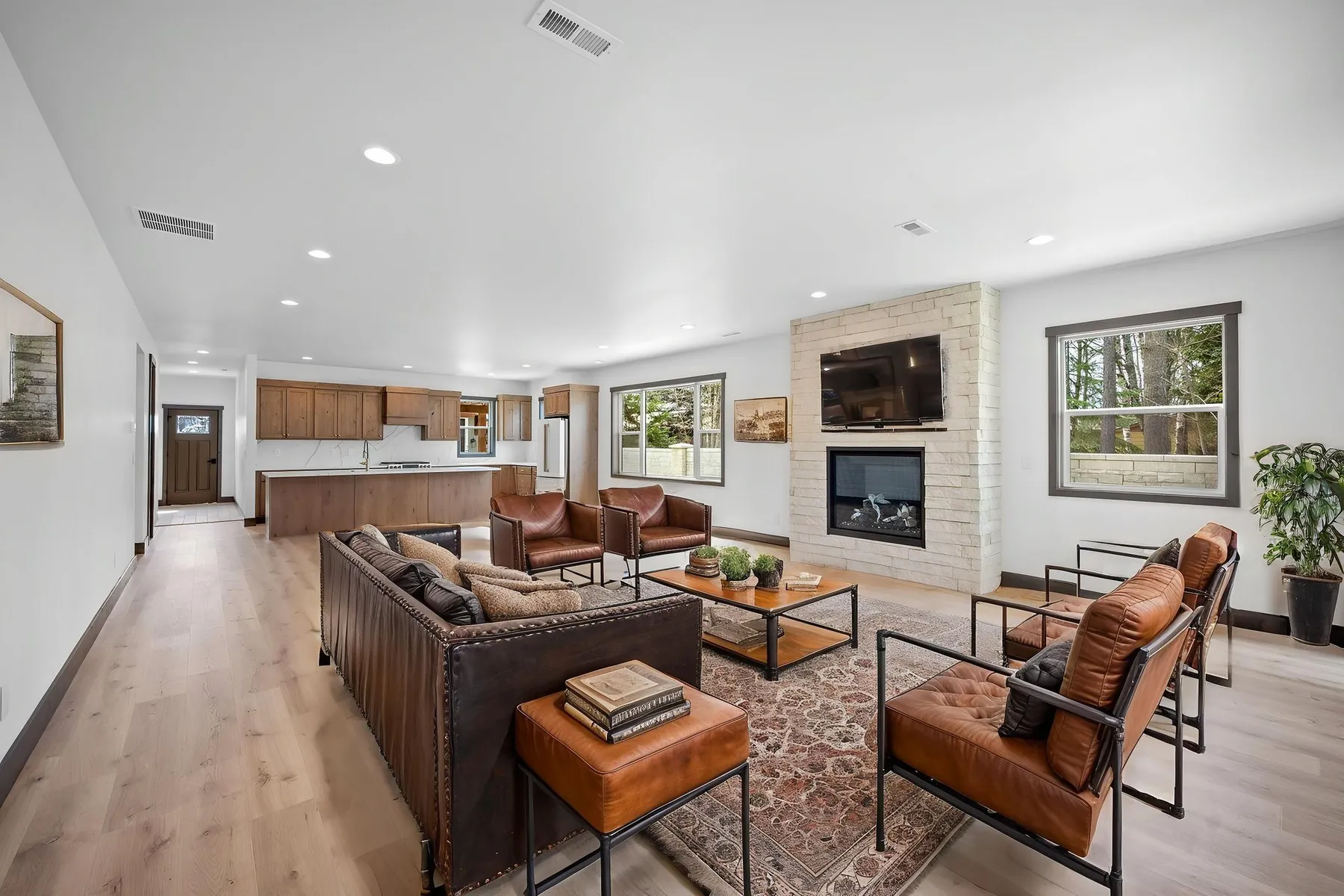 Open living room with leather seating around a stone fireplace with a mounted TV, opening onto the kitchen at the back of the house.