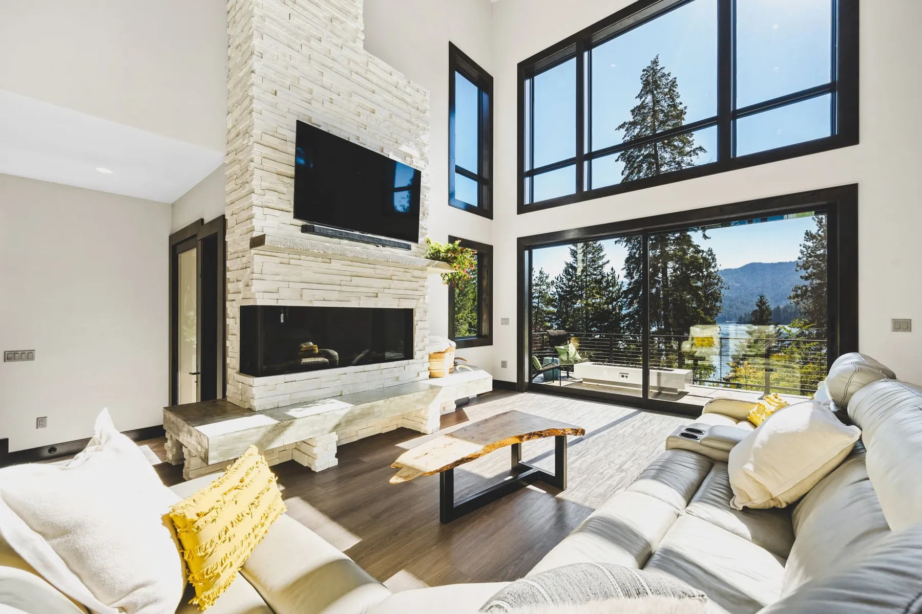 Two-story living room with a white stone fireplace, a mounted TV and floor-to-ceiling black-framed windows looking out to Hayden Lake.