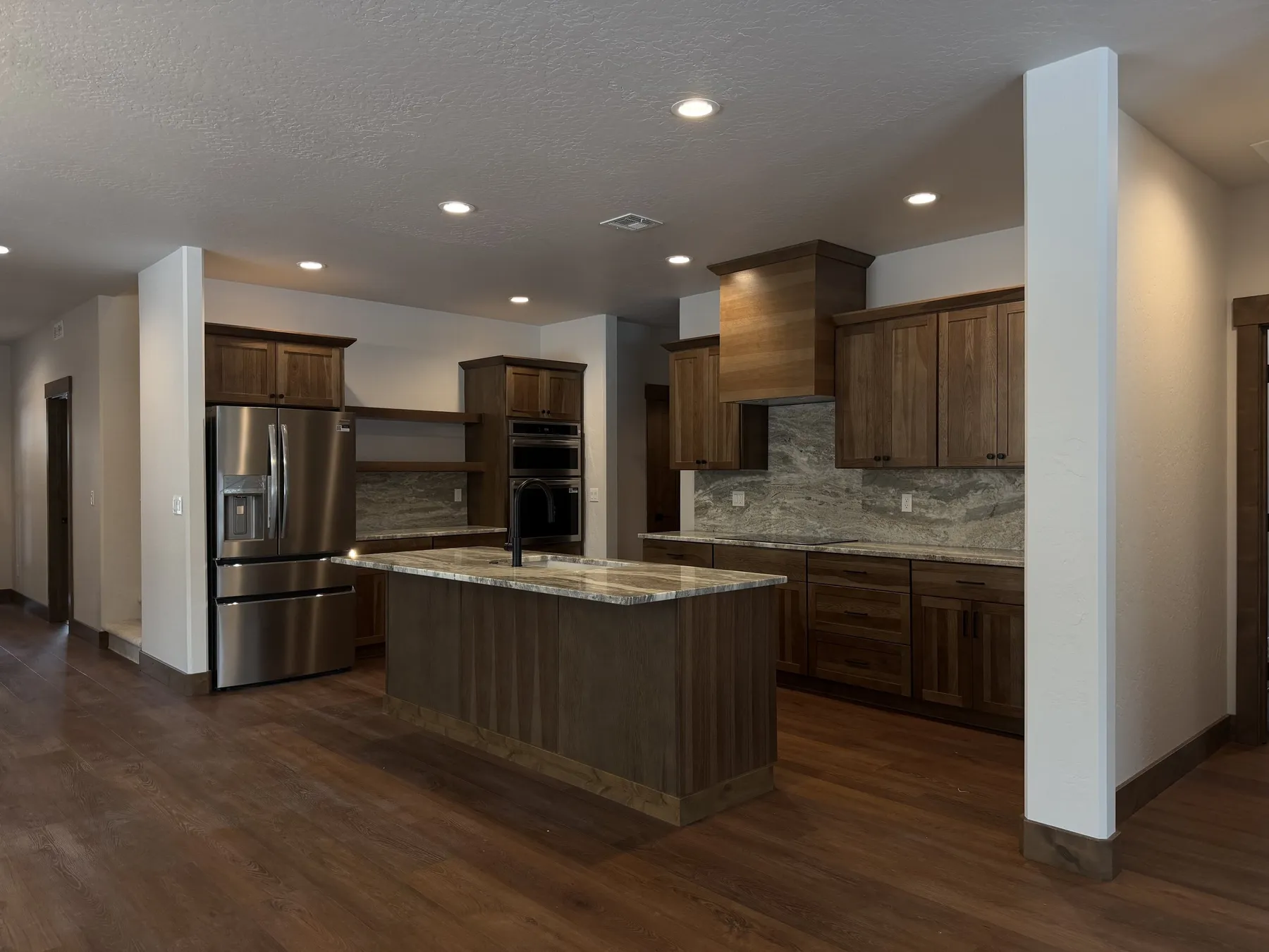 Kitchen with dark wood cabinetry, a stone-topped island, stainless double ovens and a tall wood range hood.
