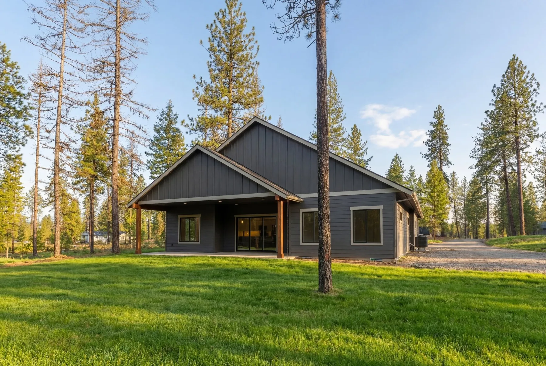 Back of the Radiant home with a covered patio on timber posts, gray siding and a lawn shaded by pines.